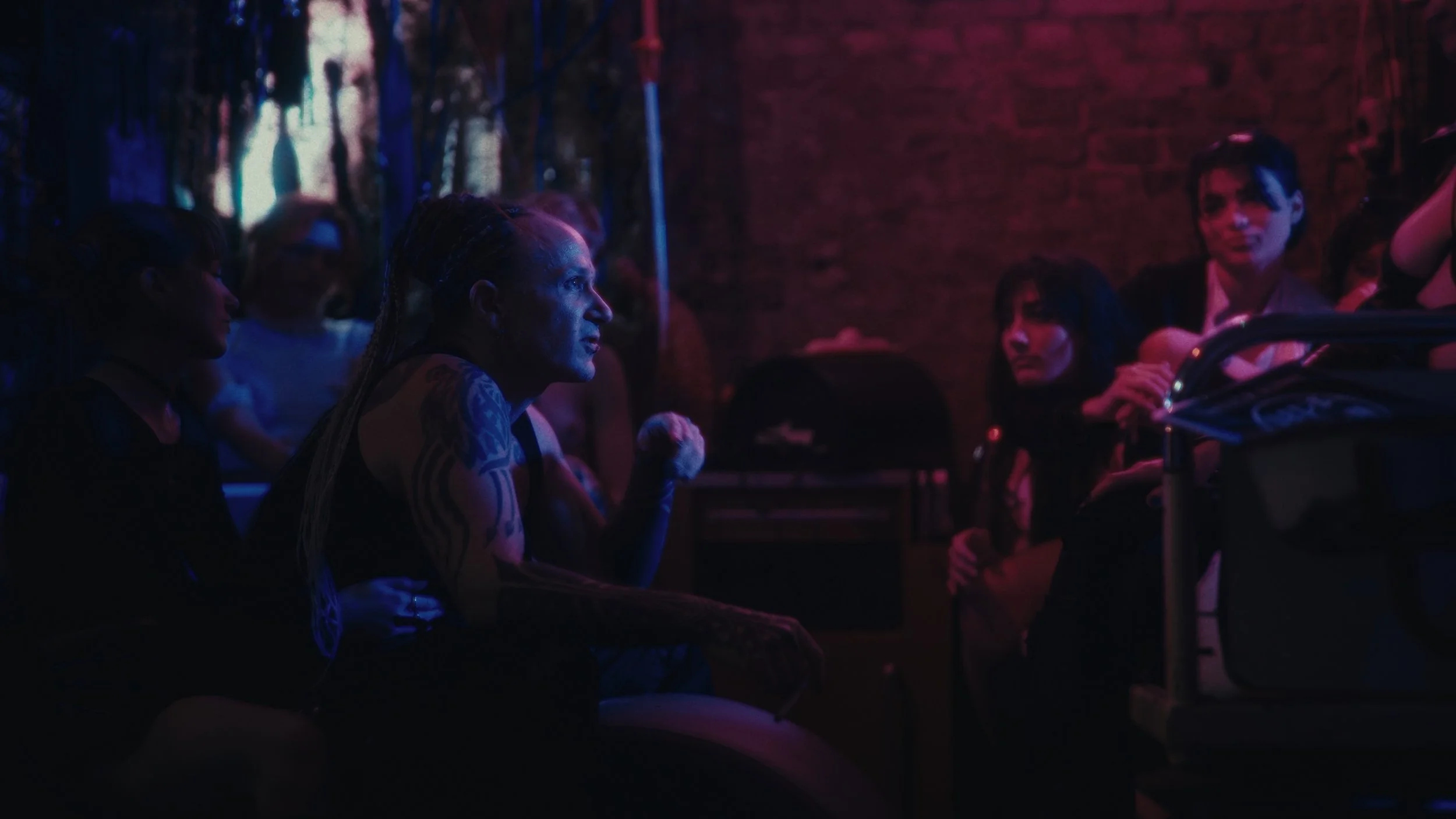 Ragazza con tatuaggi in abbigliamento nero seduta in un ambiente con luci colorate, che ascolta attentamente qualcuno durante una conversazione o presentazione.