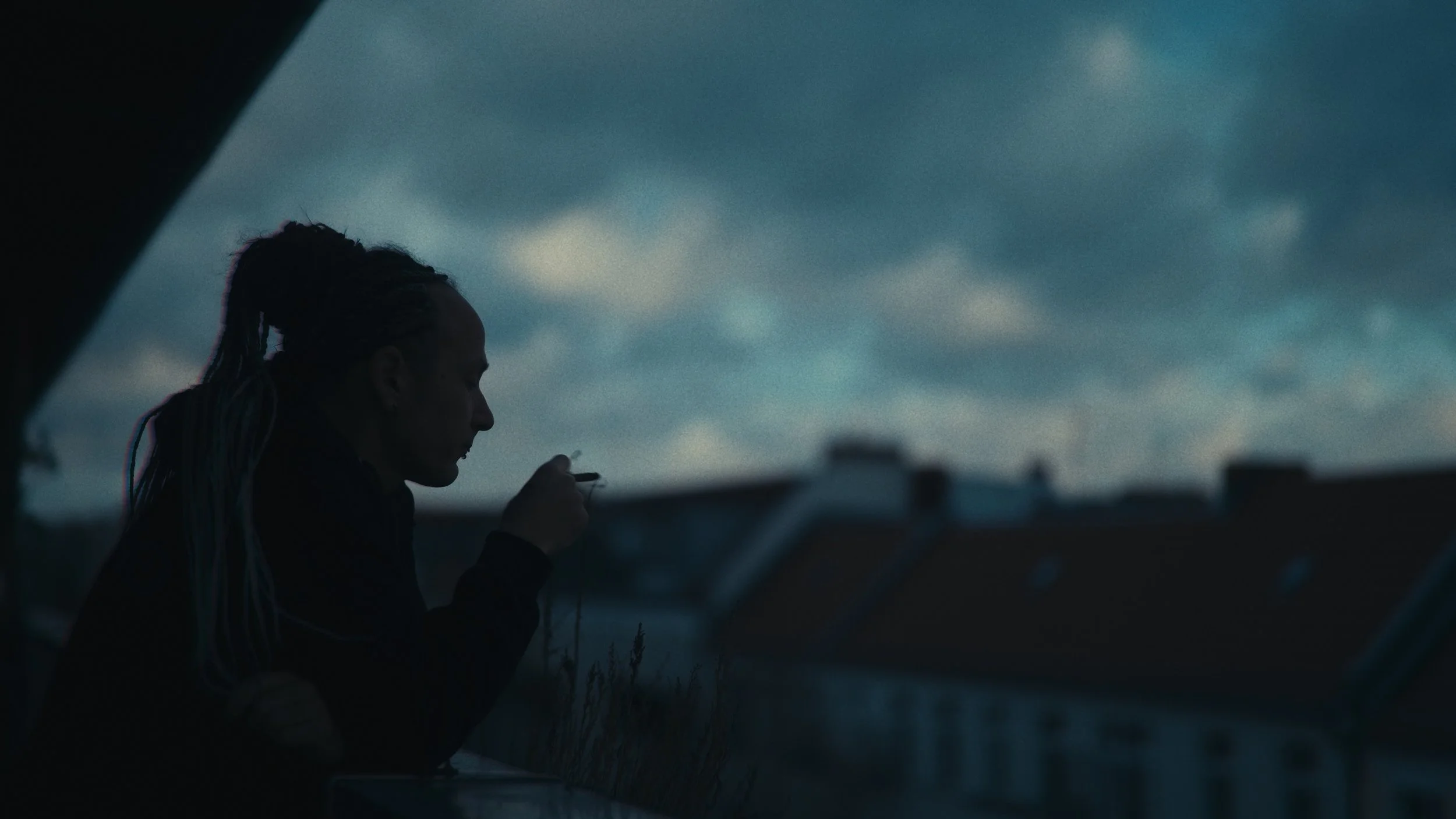 Una donna con i capelli riccioli raccolti in una coda di cavallo, seduta in un luogo all'aperto, che si dedica a fumare o a usare un oggetto con le mani, con uno sfondo di cielo nuvoloso e edifici sfocati all'orizzonte.