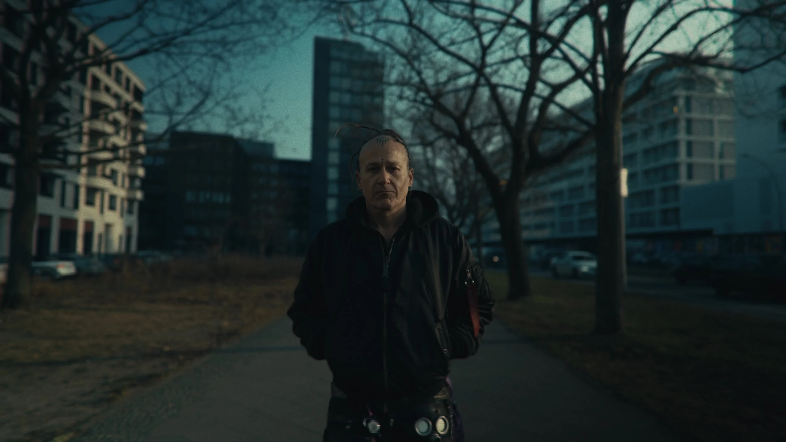 Uomo con capelli corti e tatuaggi, vestito in cappotto nero, si trova al centro di una strada urbana con edifici e alberi senza foglie sullo sfondo, in un'ambientazione serale o crepuscolare.