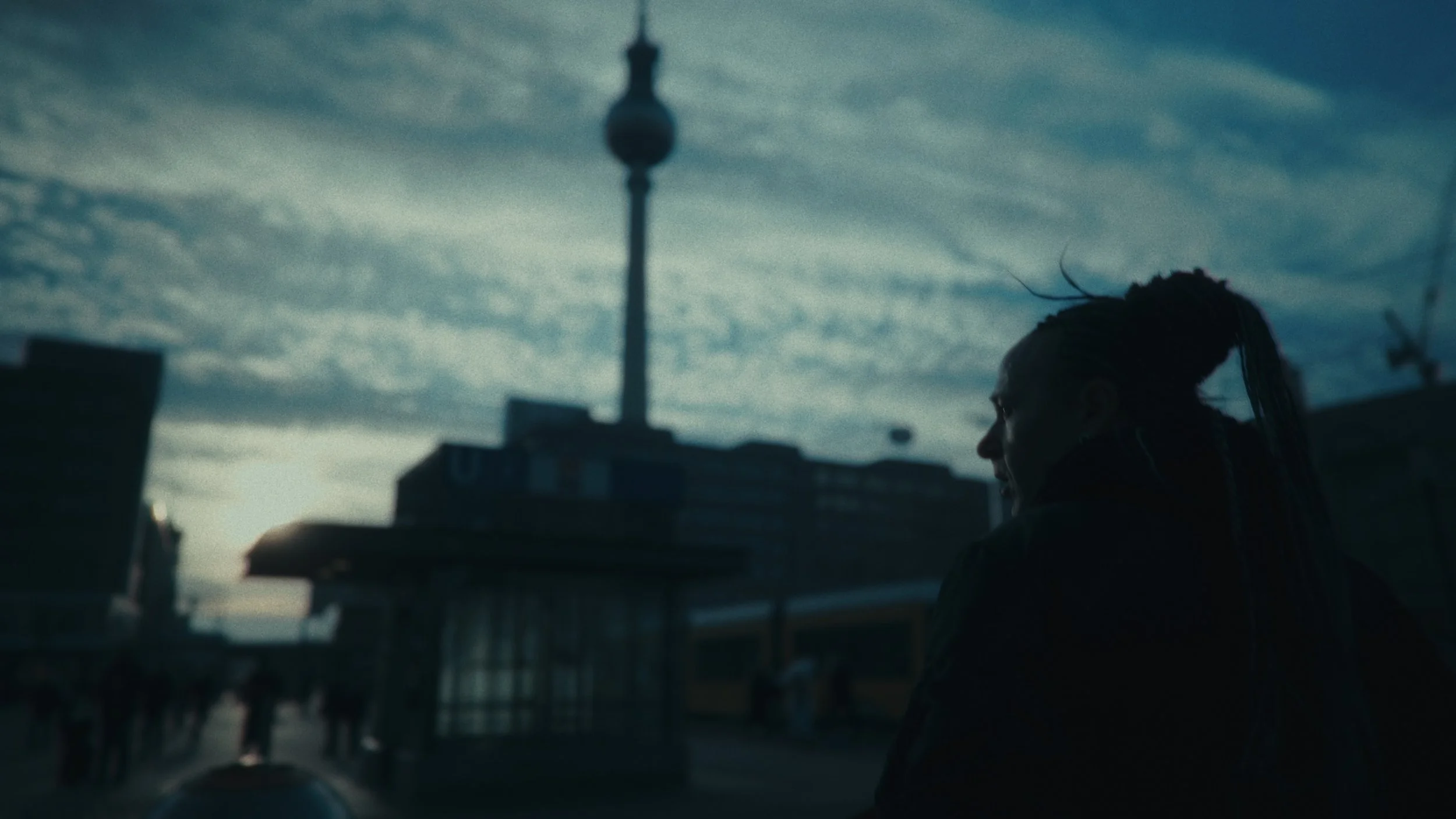 Persona con capelli in dreadlocks che guarda di lato, sullo sfondo un cielo nuvoloso e un'antenna di Berlino.