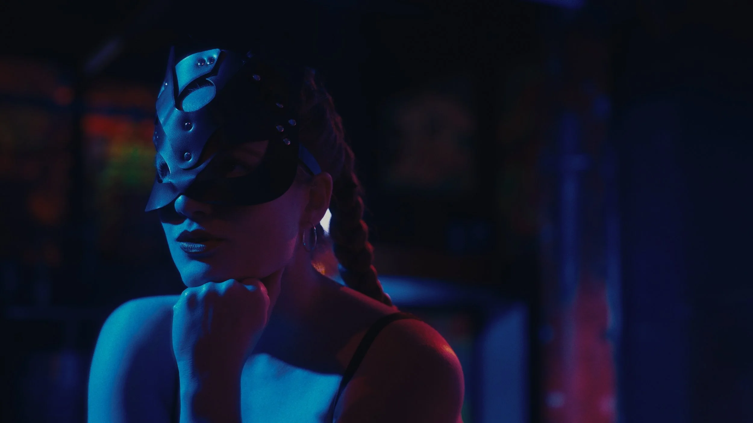 Una donna con i capelli raccolti in due trecce, indossa un cappello di maschera nera e vestiti scuri, in un ambiente con luce soffusa