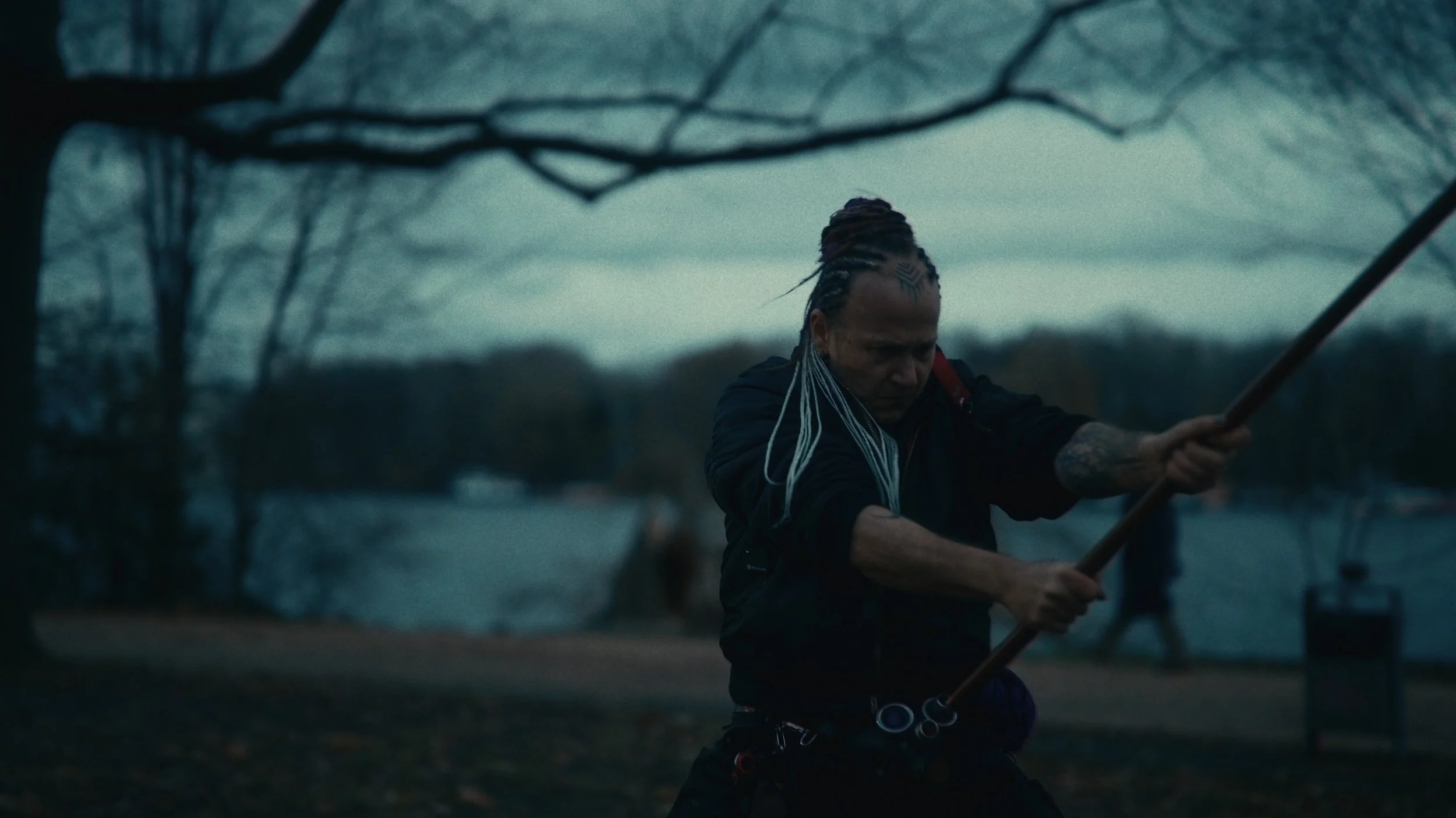 Persona che brandisce un bastone in un parco presso un lago, ambientazione crepuscolare.