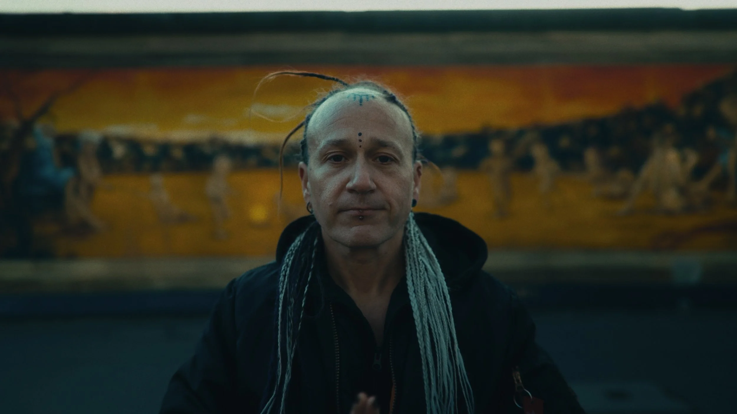 Un uomo con capelli androgeni con dimorfismo, stile punk con piercing e tatuaggi sul volto, sfondo di uno skateboard decorato con figure antropomorfe.