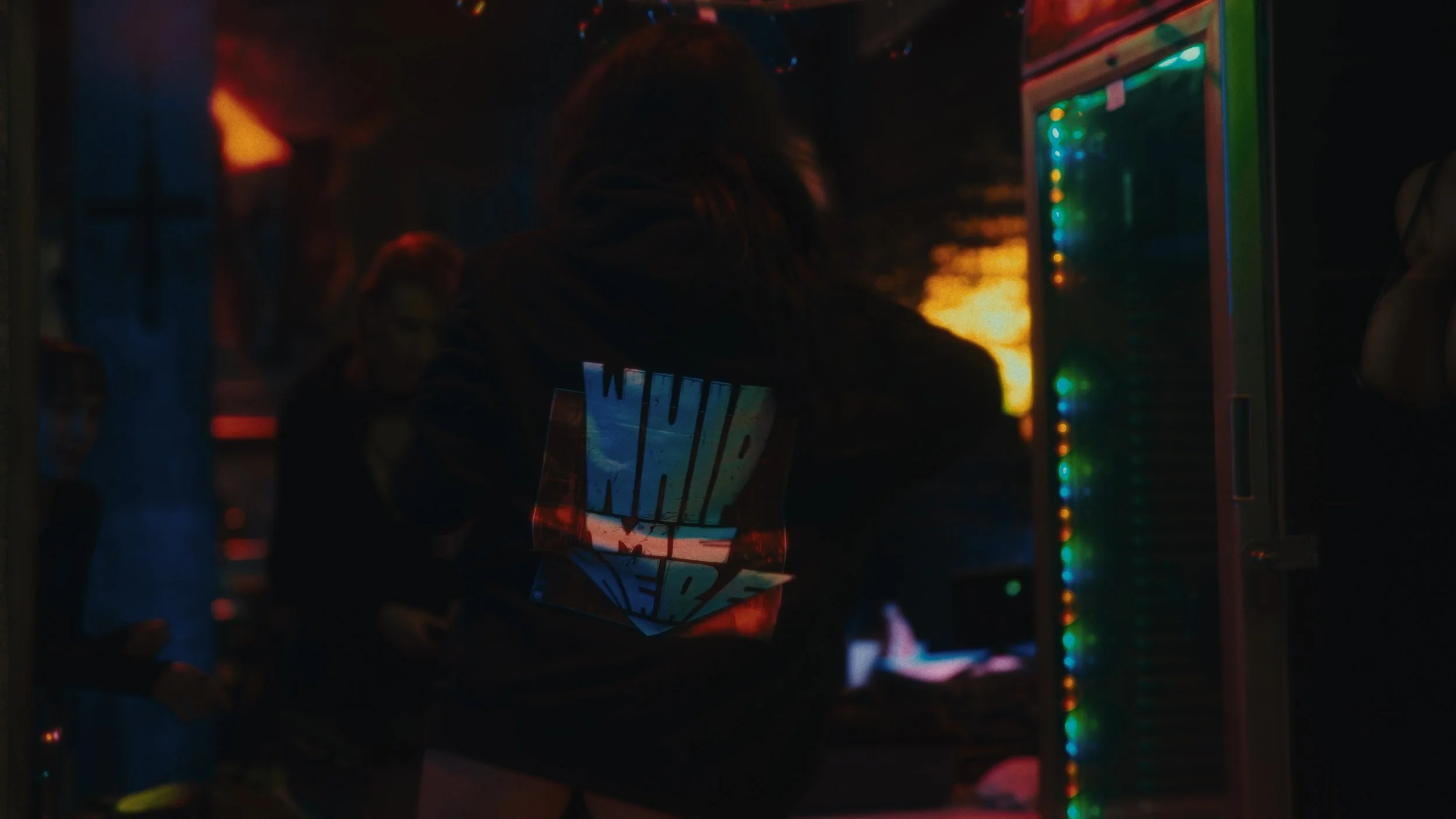 Persona in un ambiente con luci colorate, indossa felpa con scritta 'NHL Steelers' e sta davanti a una macchina di giochi arcade in ambiente oscuro.