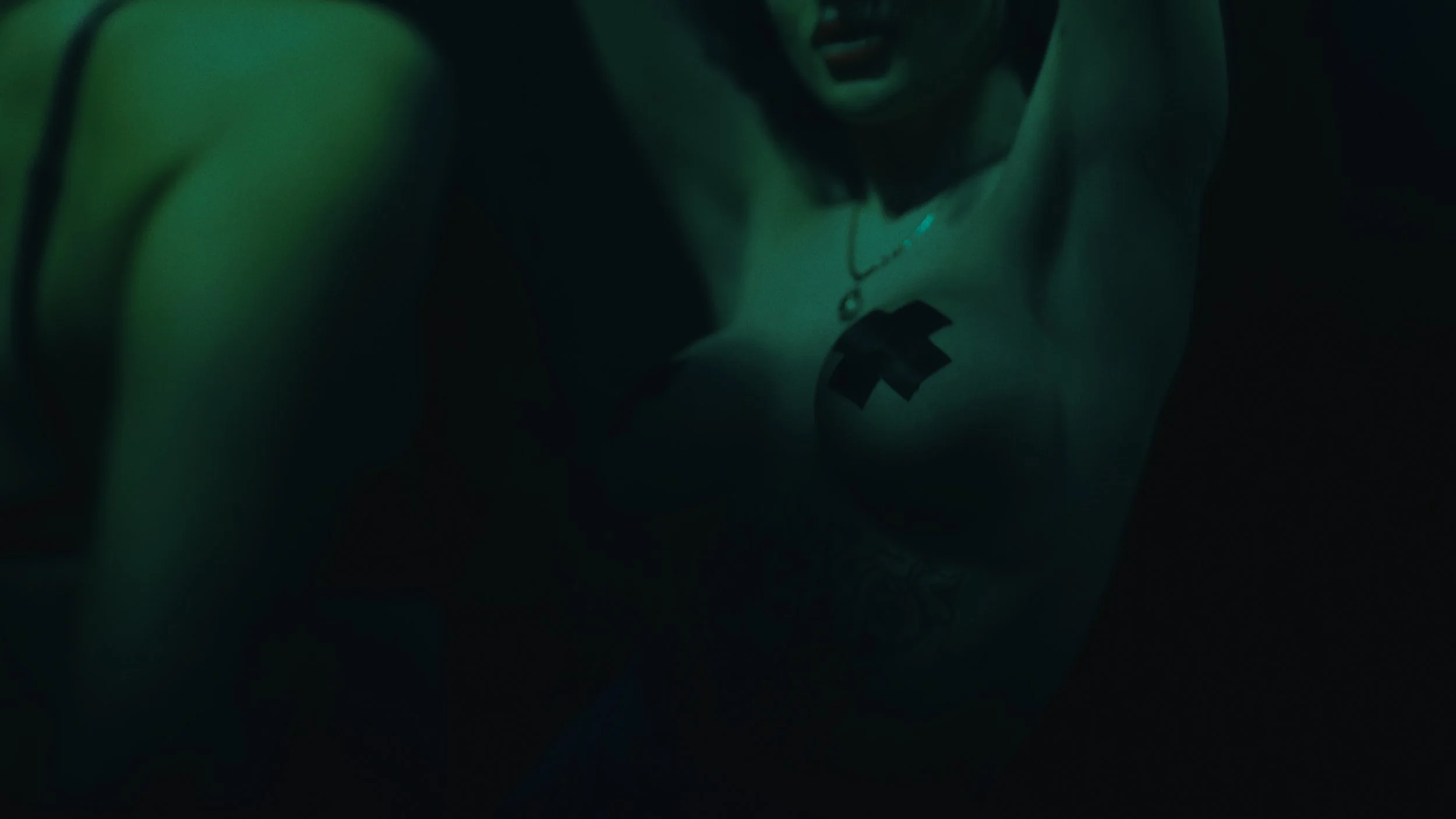 Immagine scura di una persona con pelle chiara, con tatuaggi sulla spalla e un vestito senza maniche, in un'illuminazione verde, con un'espressione aggressiva.