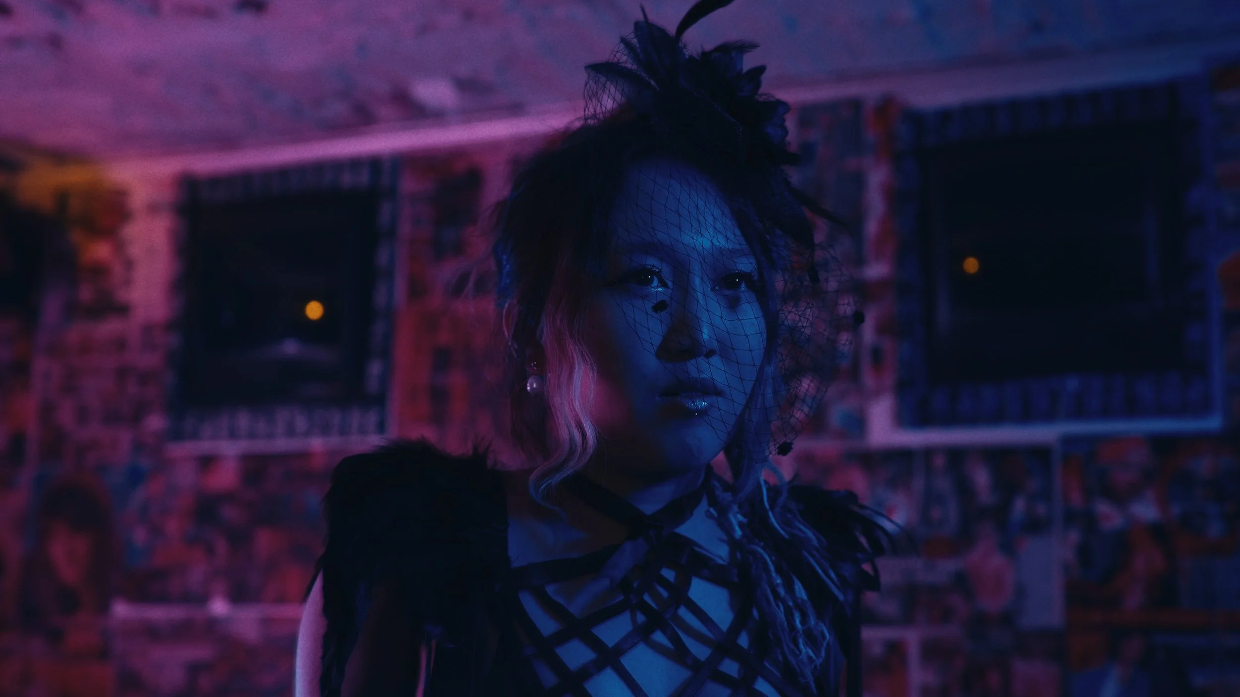 Ragazza in un ambiente con luci colorate blu e viola, indossa un'acconciatura elaborata e un vestito nero con dettagli a rete.