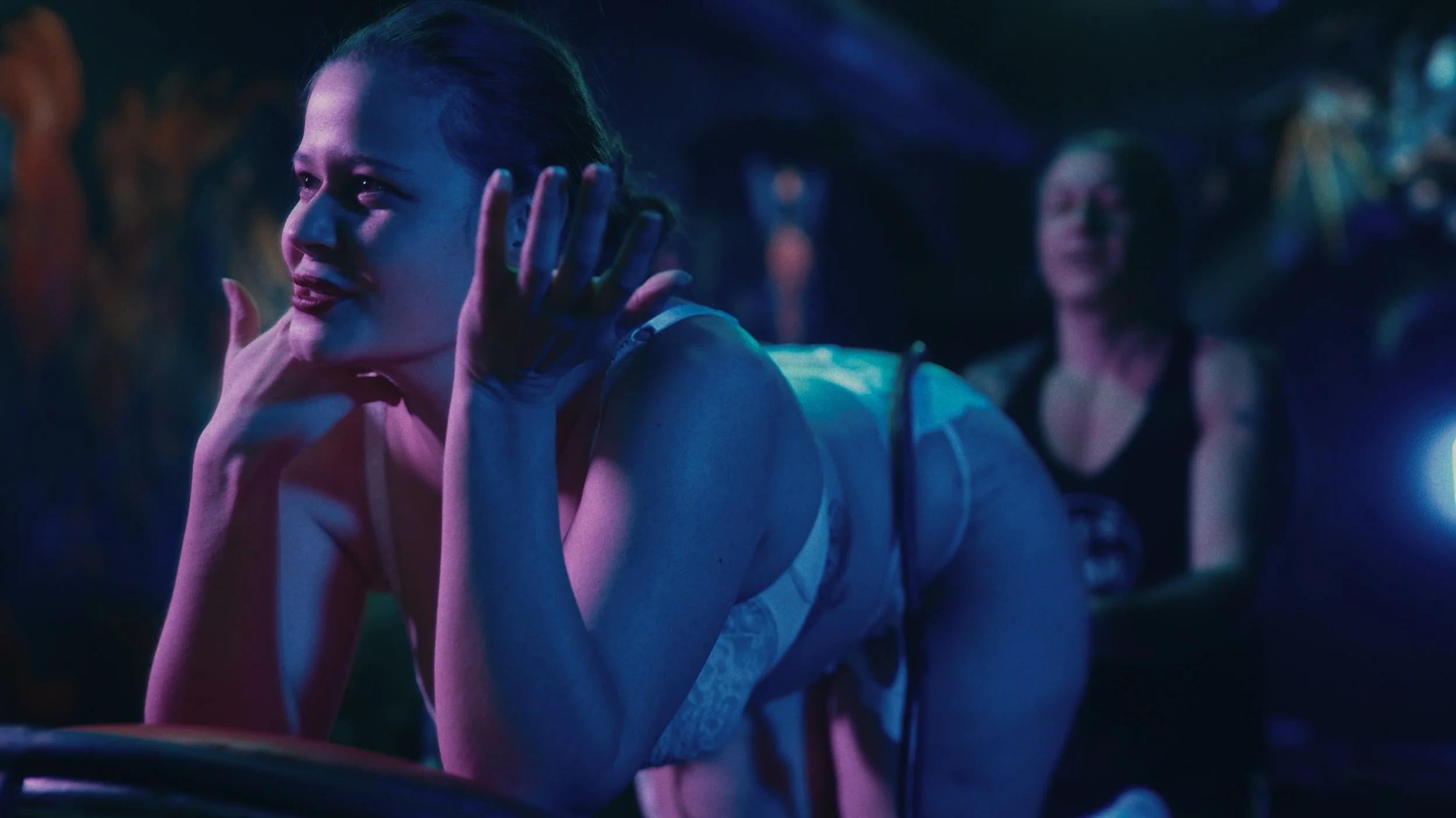 Due donne in un ambiente colorato con luci blu e viola, una in primo piano che sorride e nega con la testa, l'altra sullo sfondo che si rilassa.
