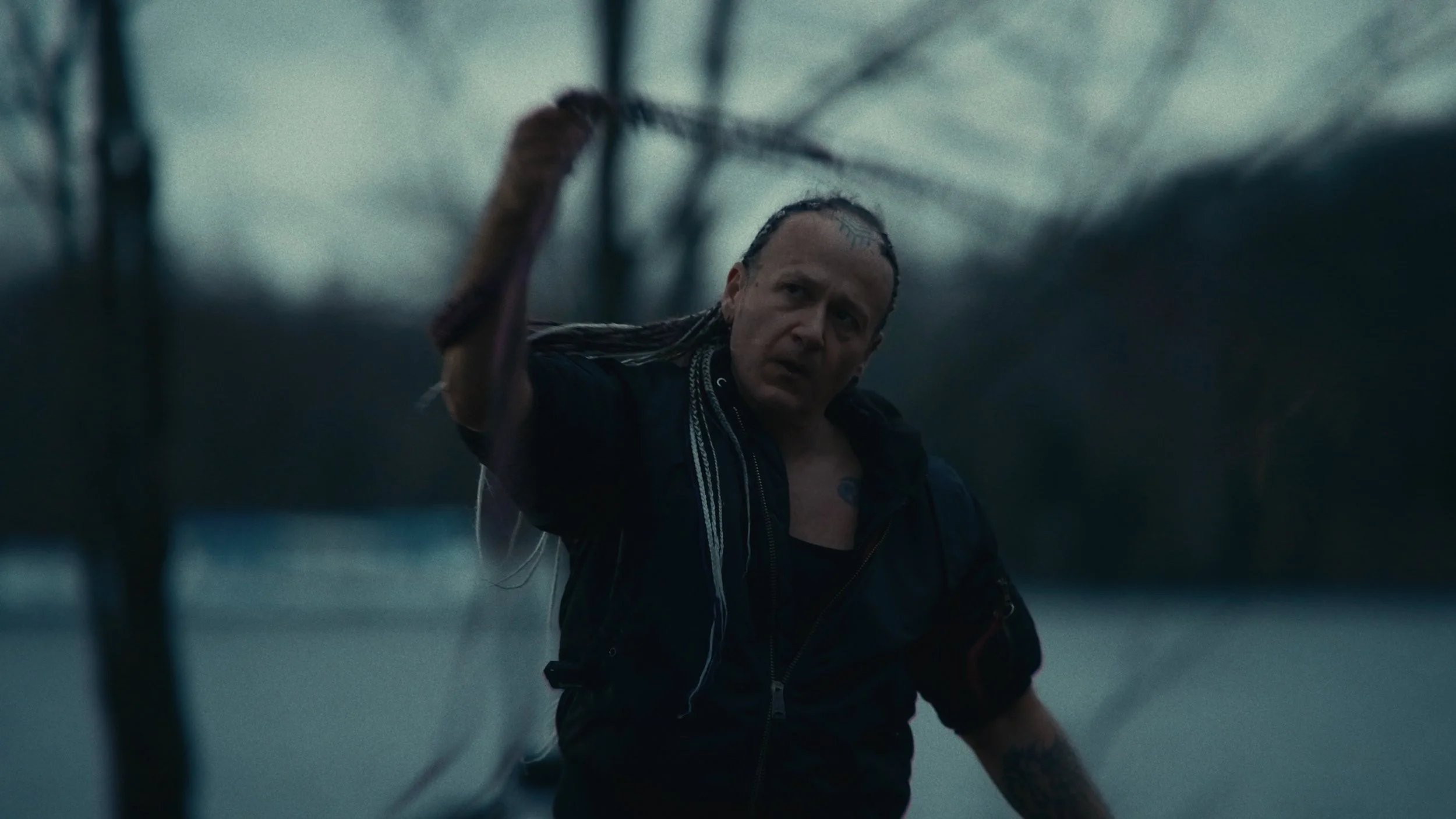 Un uomo con capelli lunghi e tatuaggi solleva un ramo di un albero vicino a un lago. L'ambiente è durante il crepuscolo o l'alba, con un cielo nuvoloso e un'atmosfera malinconica.