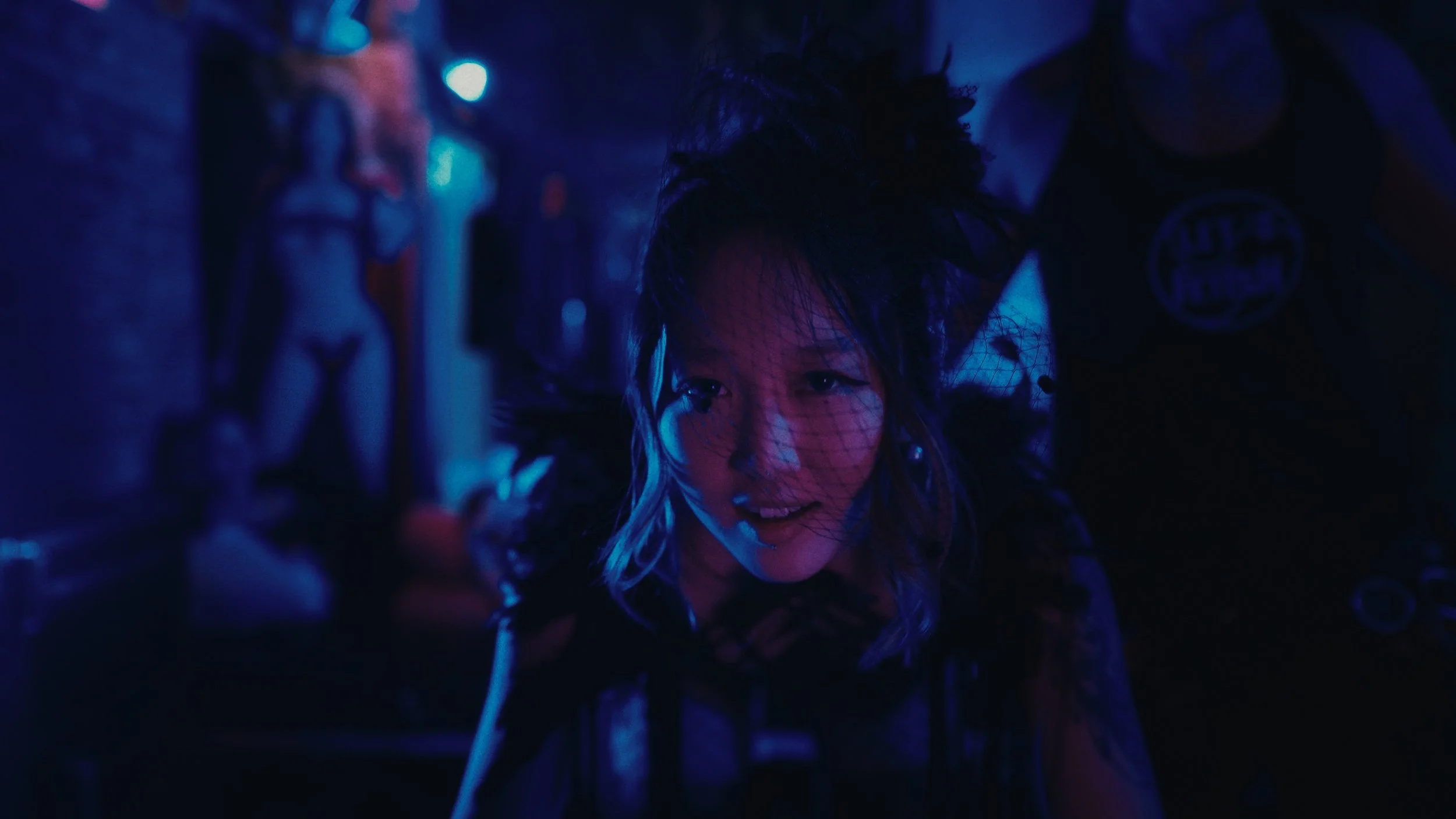 Una donna con trucco teatrale, indossa un cappello nero e una rete sul viso, sorride in un ambiente con illuminazione blu e viola, con un manichino in costume sullo sfondo.