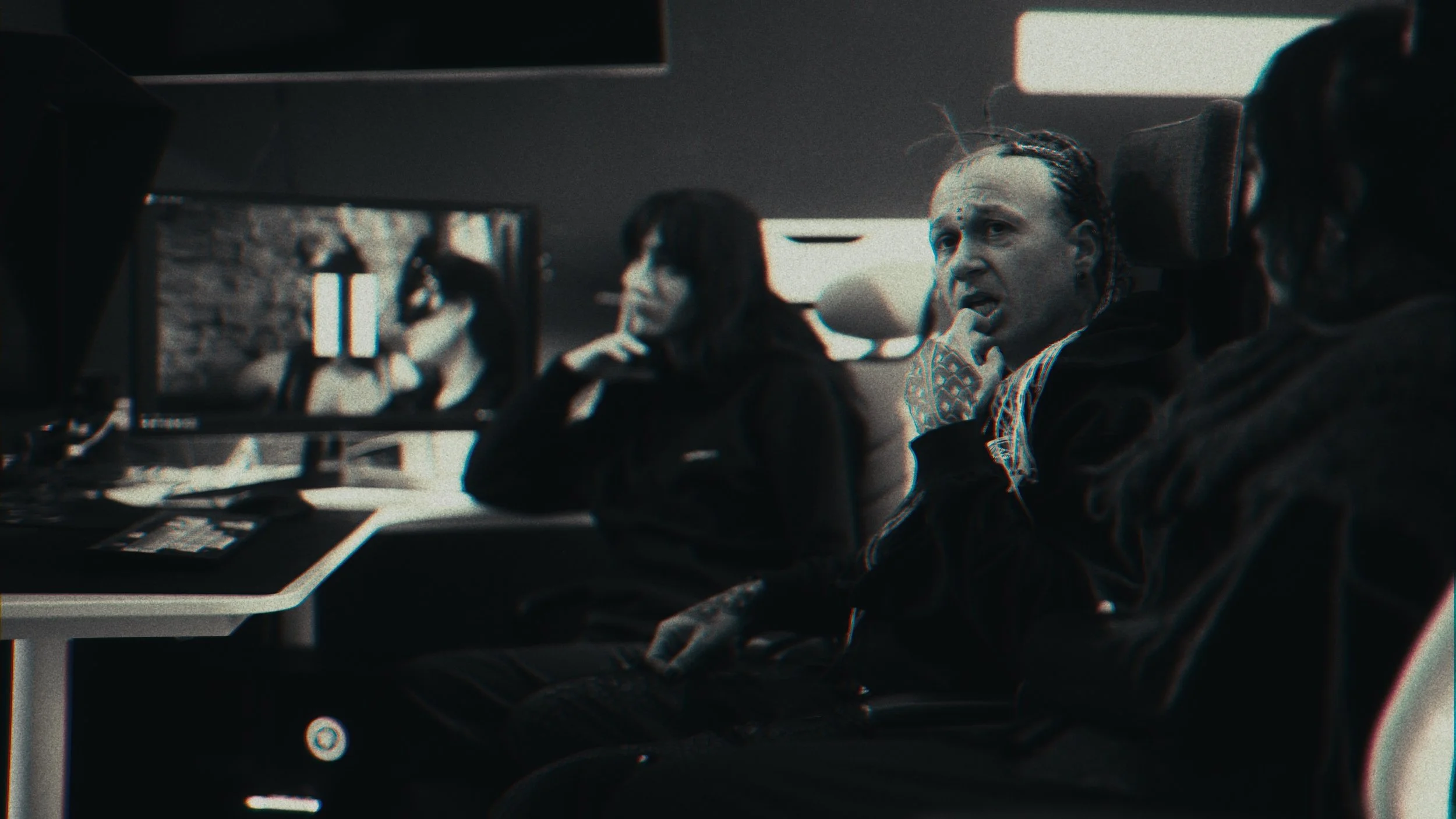 Tre persone sedute in una stanza buia, una con capelli riccioli e una con treccine, stanno guardando uno schermo di computer, visibile anche in una monitor che mostra le stesse persone.