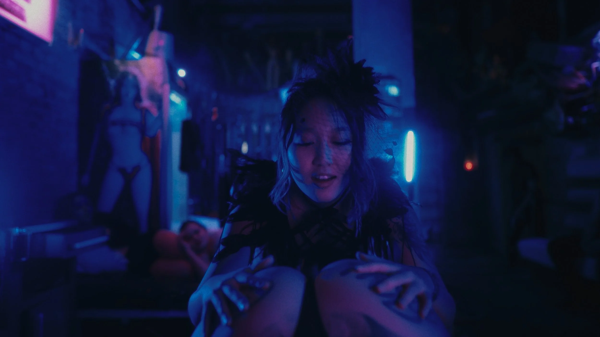 Una donna con abbigliamento dark e acconciatura originale si trova in un ambiente oscuro illuminato da luci blu e viola, con decorazioni suggestive sullo sfondo.