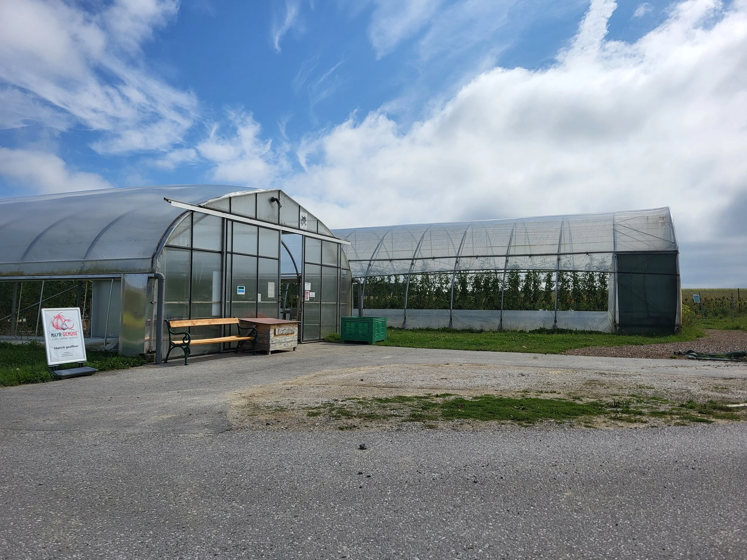 Ein Gewächshaus in einem Feld mit einem freien Platz davor. Der Himmel ist bewölkt mit blauen Bereichen.