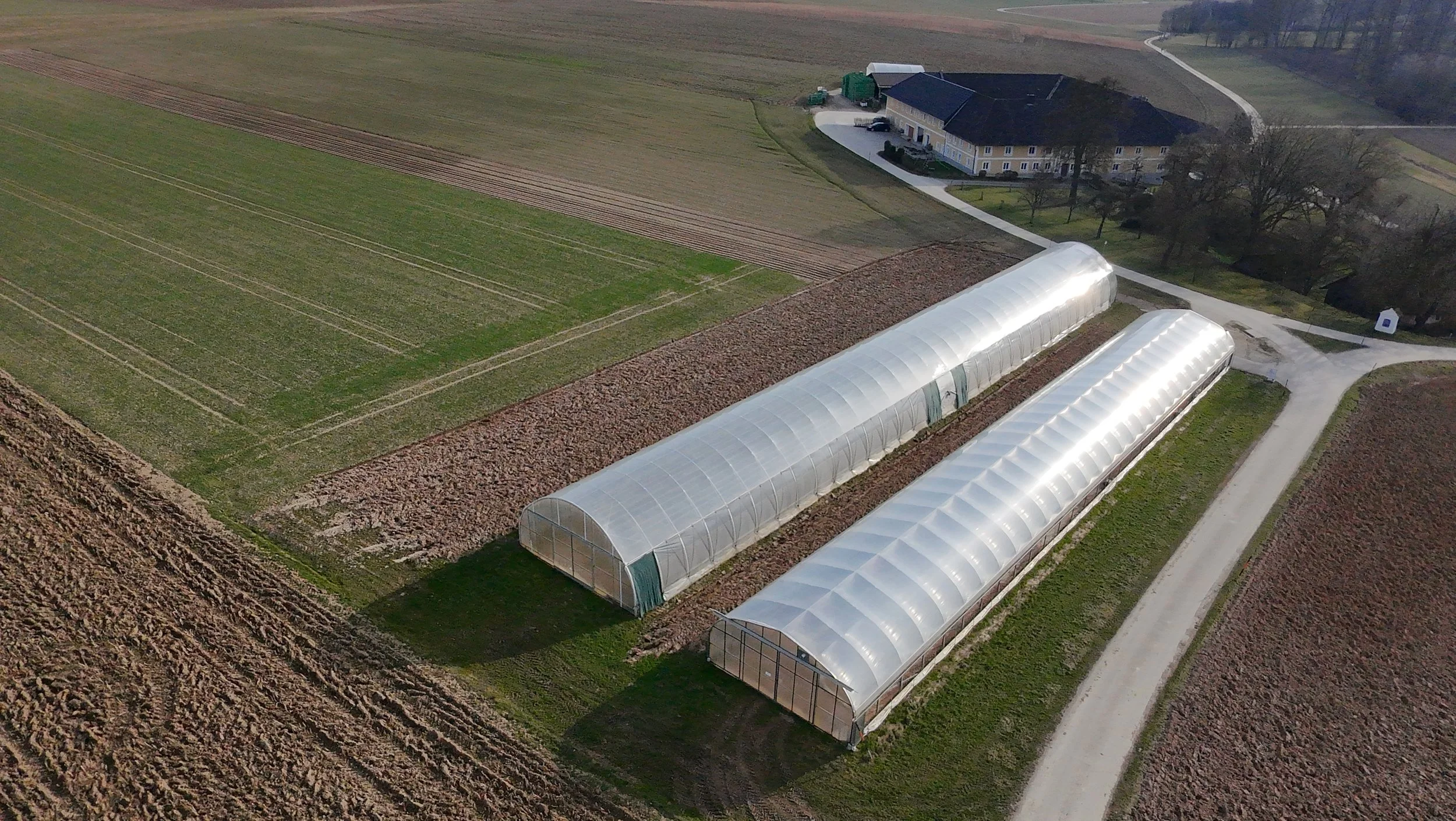 Luftaufnahme eines landwirtschaftlichen Betriebs mit zwei gewölbekarierten Gewächshäusern, Feldern und einem großen ländlichen Gebäude mit mehreren Eingängen, umgeben von Bäumen und Wegen.