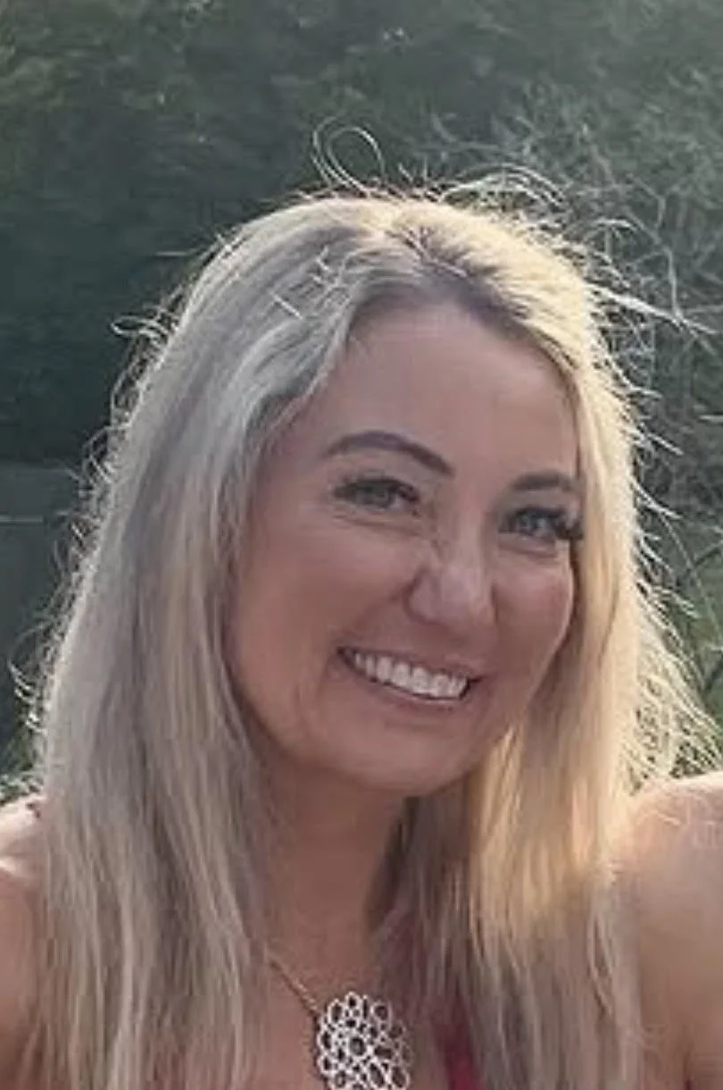 A woman with blonde, wavy hair and blue eyes, wearing a black sleeveless top and a silver necklace with a circular pendant, sitting at a table with a notebook, smiling at the camera.