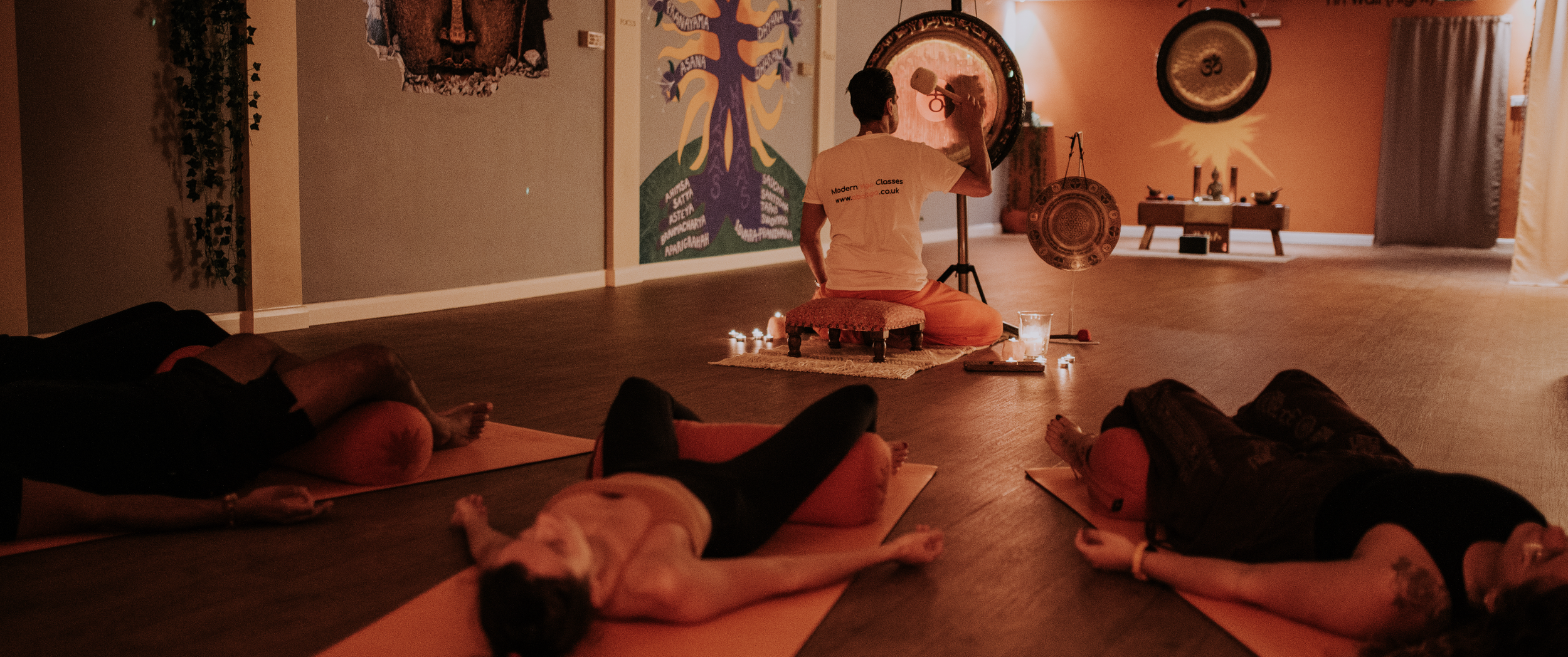 A person leading a yoga or meditation class in a warm, dimly lit room with three students lying on mats and a dog resting on one of the mats.