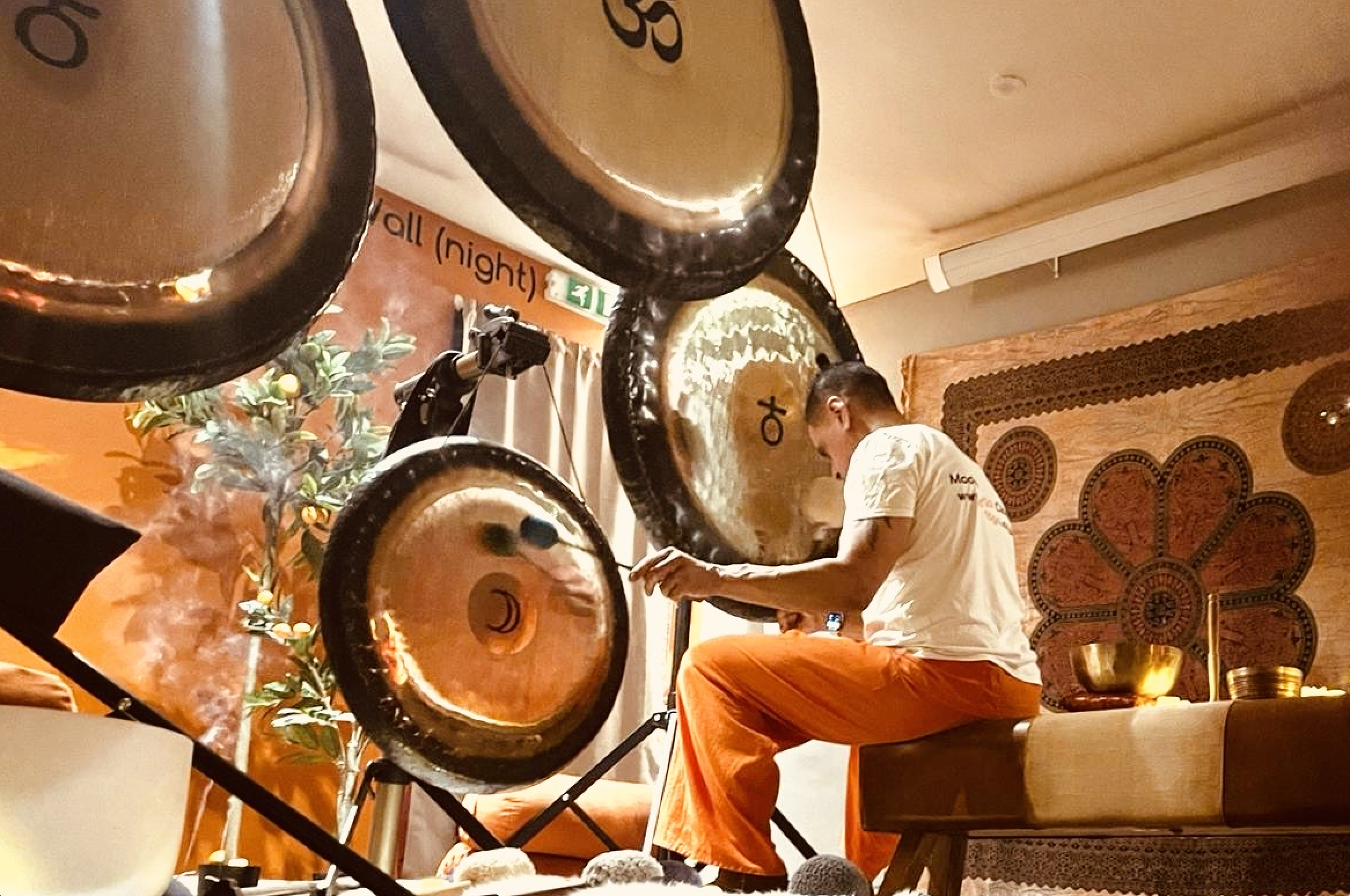 Chris Abay sitting on a low bench playing a gong, surrounded by several large hanging gongs in a room with orange and brown decor, including wall art and a decorated wall panel.