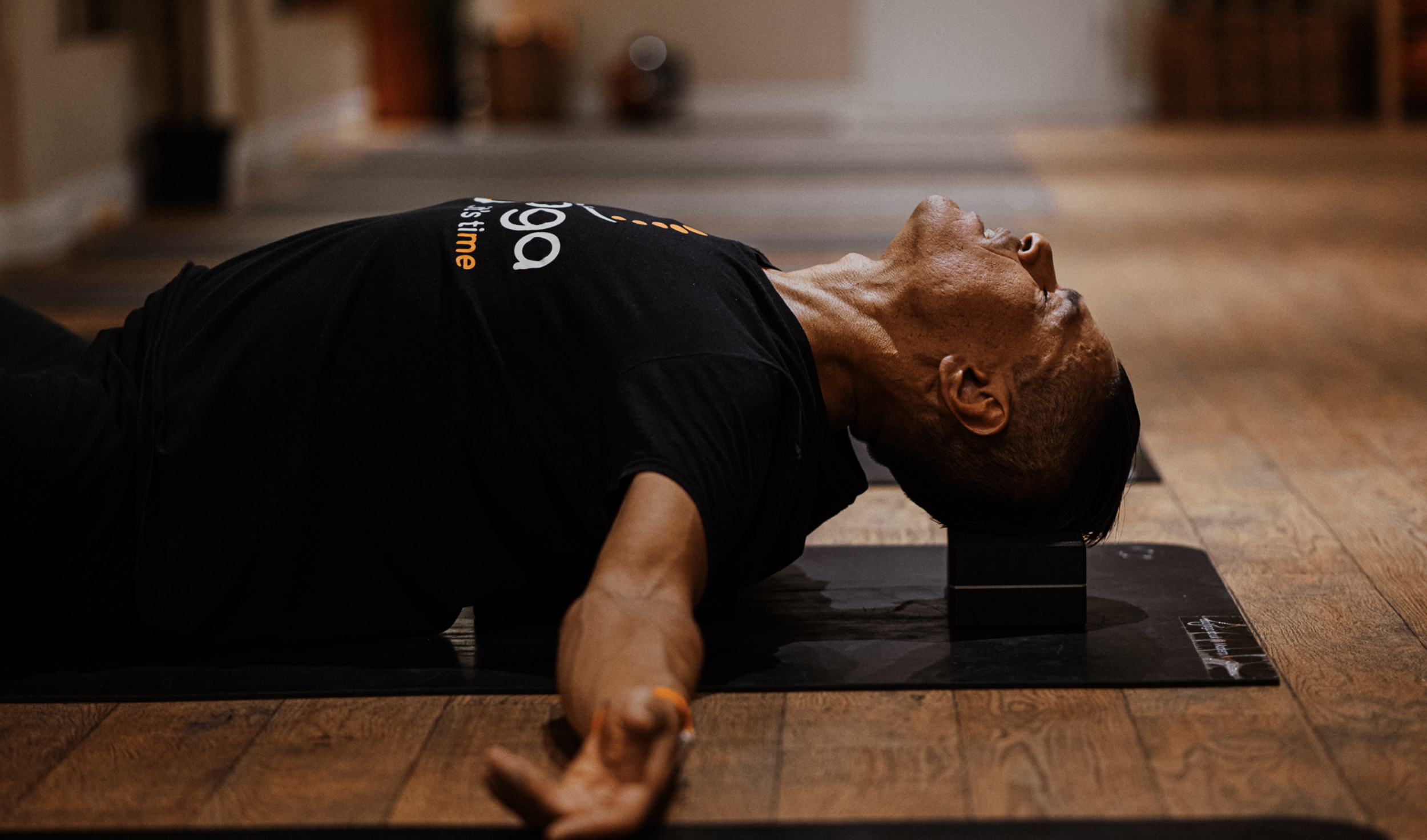 An older man doing yoga on a mat indoors, lying on his back with his head supported by a small bolster.
