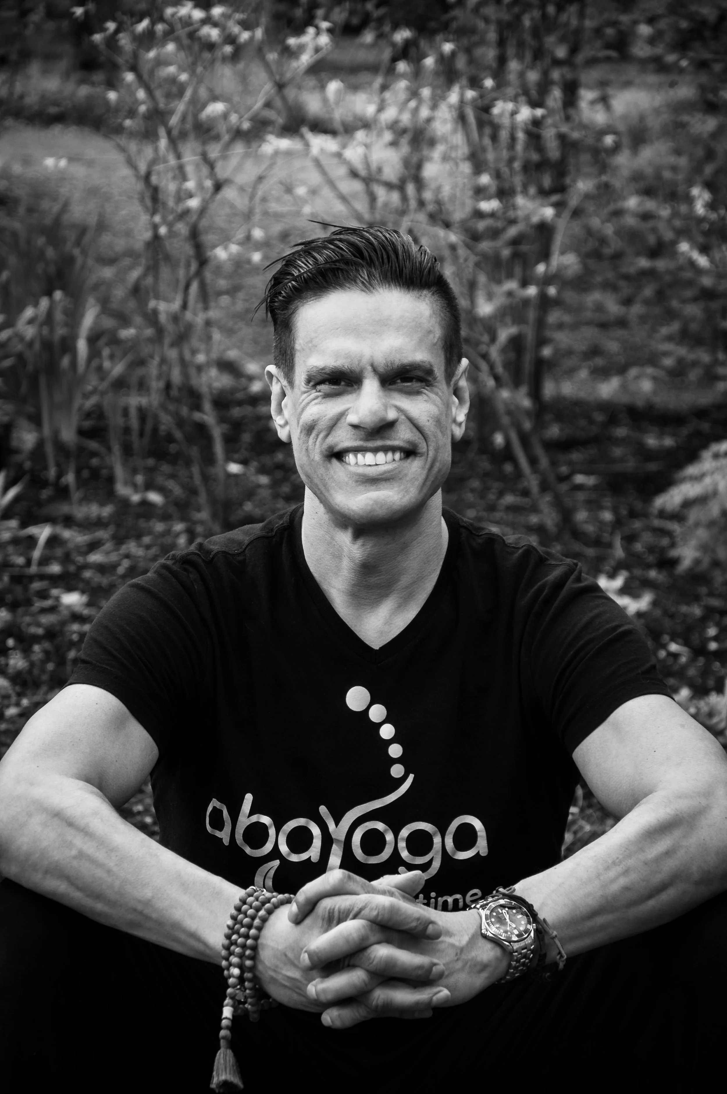 Smiling Yin Yoga Teacher with short, styled hair sitting outdoors in a garden, wearing a T-shirt with the logo 'abayoGa', a wristwatch, and beaded bracelets.