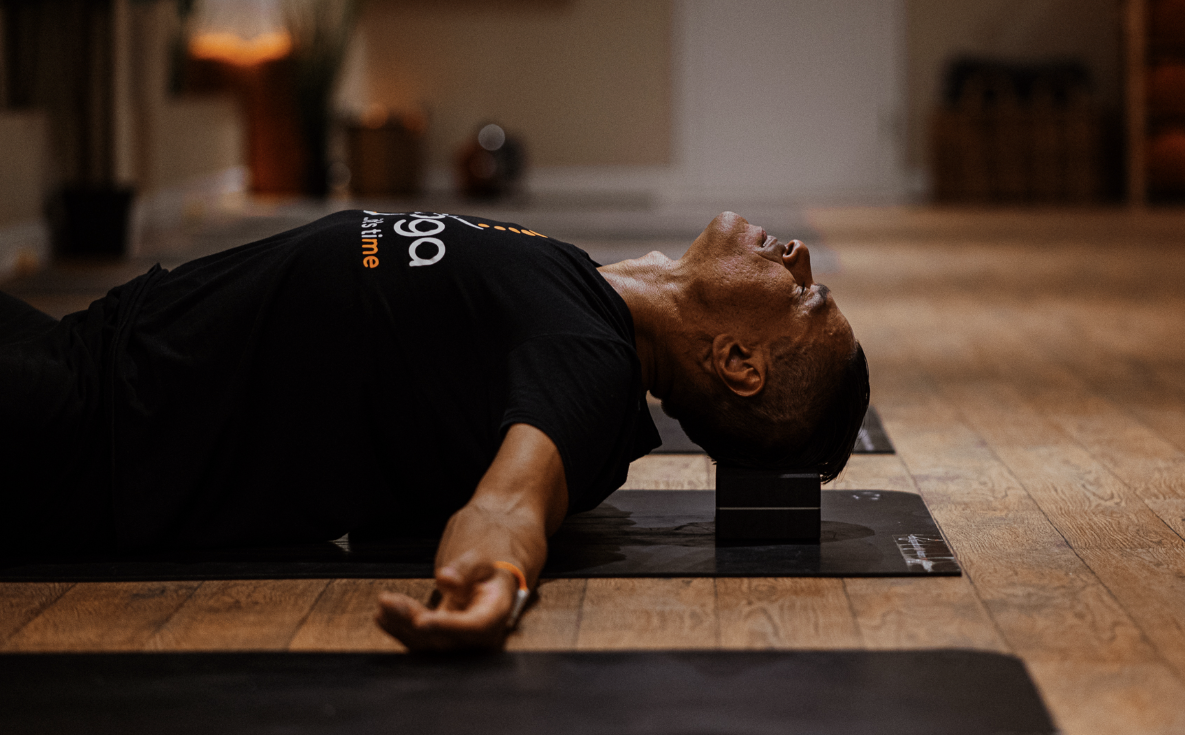 Chris Abay practicing yoga nidra indoors, lying on his back with a yoga block under his neck, arms relaxed at his sides, eyes closed, on a black yoga mat.