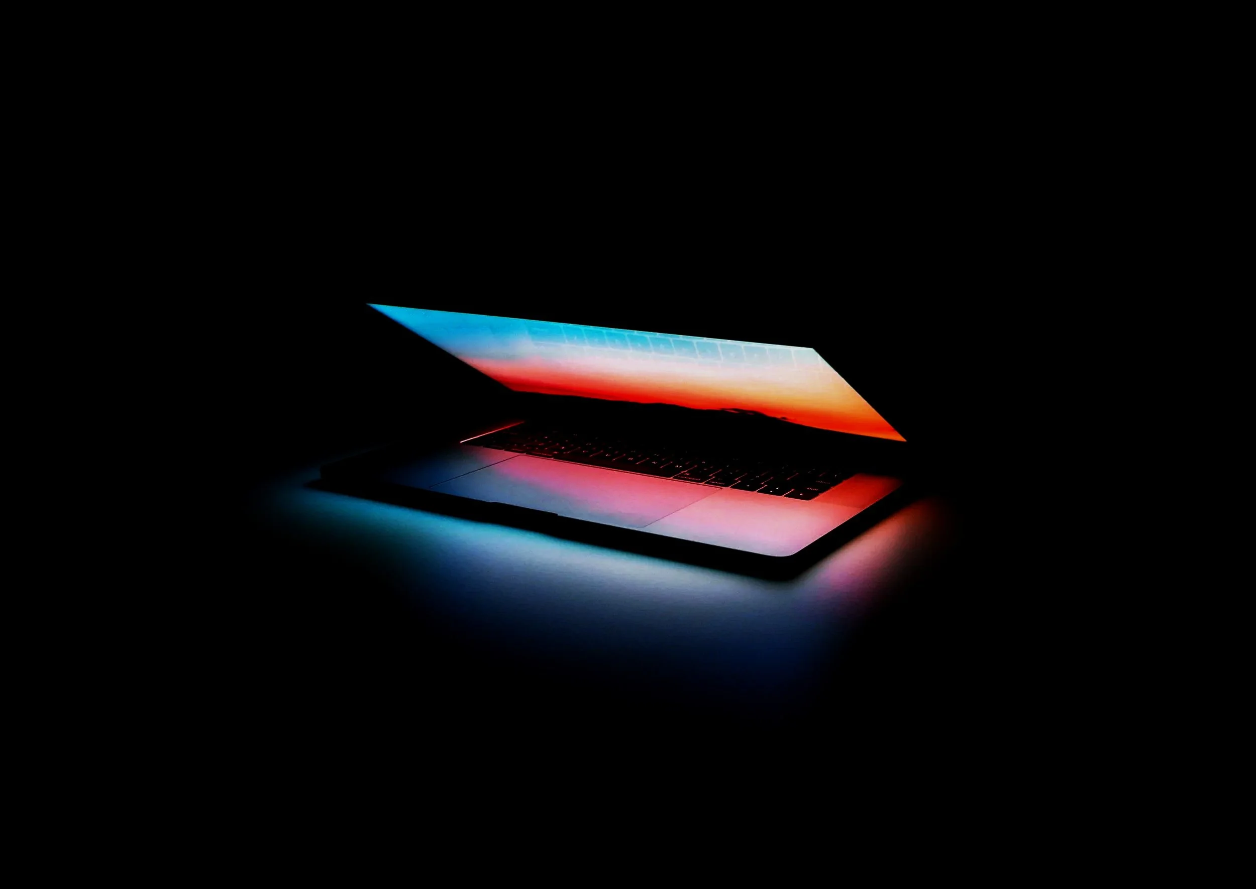 A slim laptop with a colorful sunset or sunrise image on the screen, glowing softly in a dark environment.