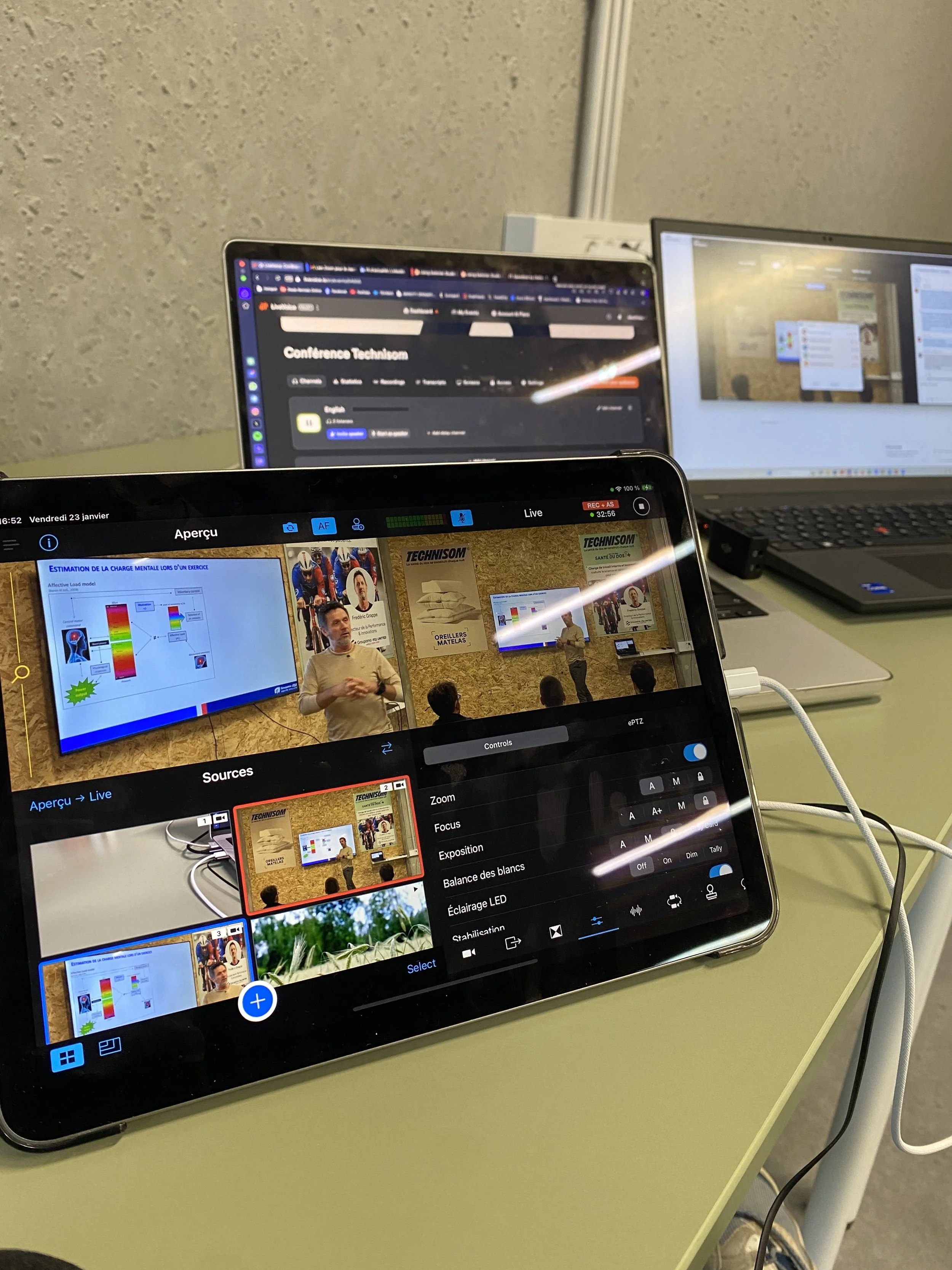 Une salle de conférence avec plusieurs ordinateurs portables sur une table, un mur en plâtre en arrière-plan et une présentation projetée sur un écran mural montrant un homme parlant lors d'une conférence.