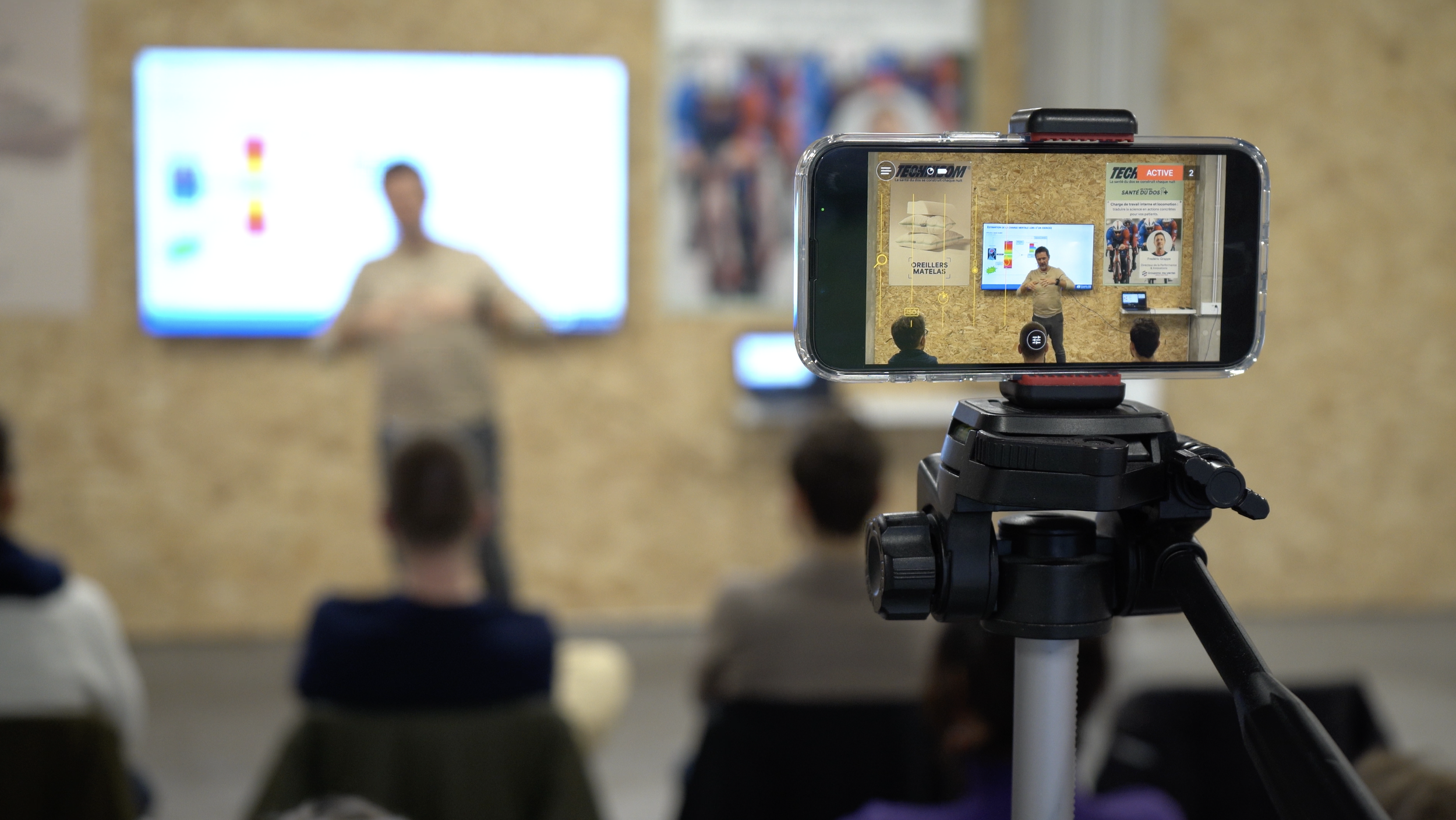 Une caméra filme une présentation dans une salle avec un écran display indiquant une personne qui parle. Le présentateur se tient devant un mur en panneau de particules avec des affiches, et il y a des personnes assises dans l'audience.