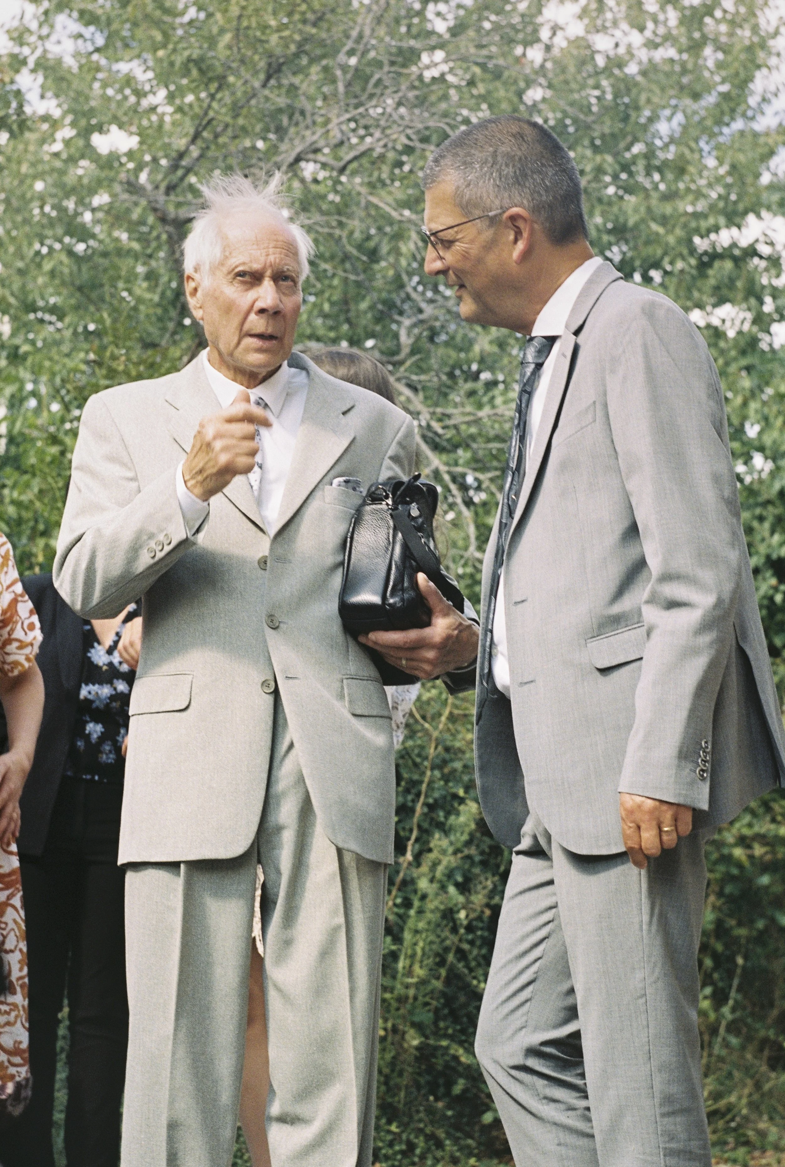 photographie de Mariage à l'argentique Martin Michel.Deux hommes en costume discutent lors d'une cérémonie en plein air, l'un d'eux porte un costume clair avec une cravate, l'autre un costume gris avec une cravate à motif.