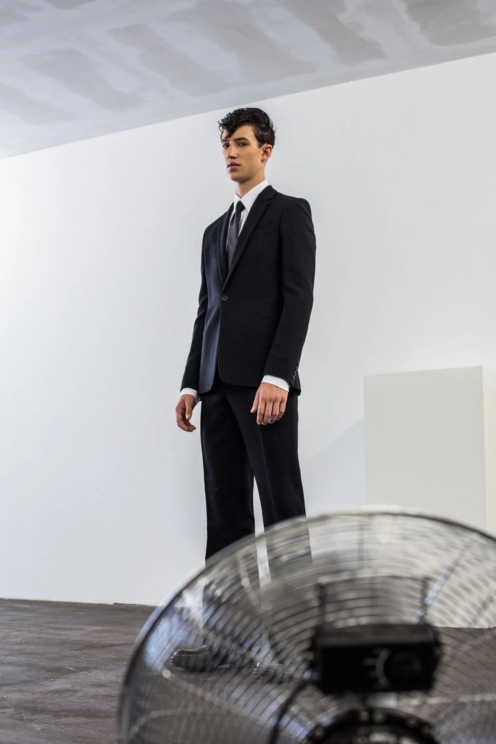 A young man in a black suit and tie standing in a minimalist, white room, with an industrial fan in the foreground.