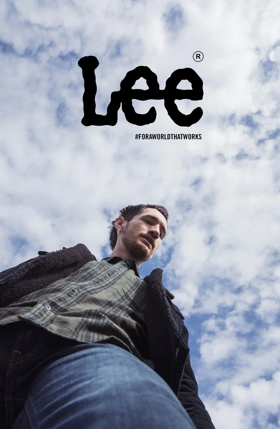 Young man looking down at the camera against a partly cloudy sky, with a large 'Lee' logo and hashtag '#foraworldthatworks' in the sky.
