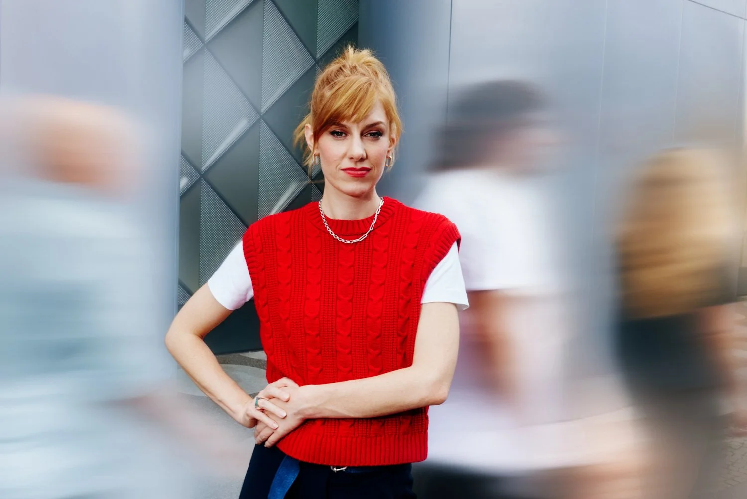 A red-haired woman in a red sweater vest and white t-shirt is standing in an urban setting with metallic walls. She is wearing silver jewelry and has her hands clasped in front of her, looking directly at the camera.