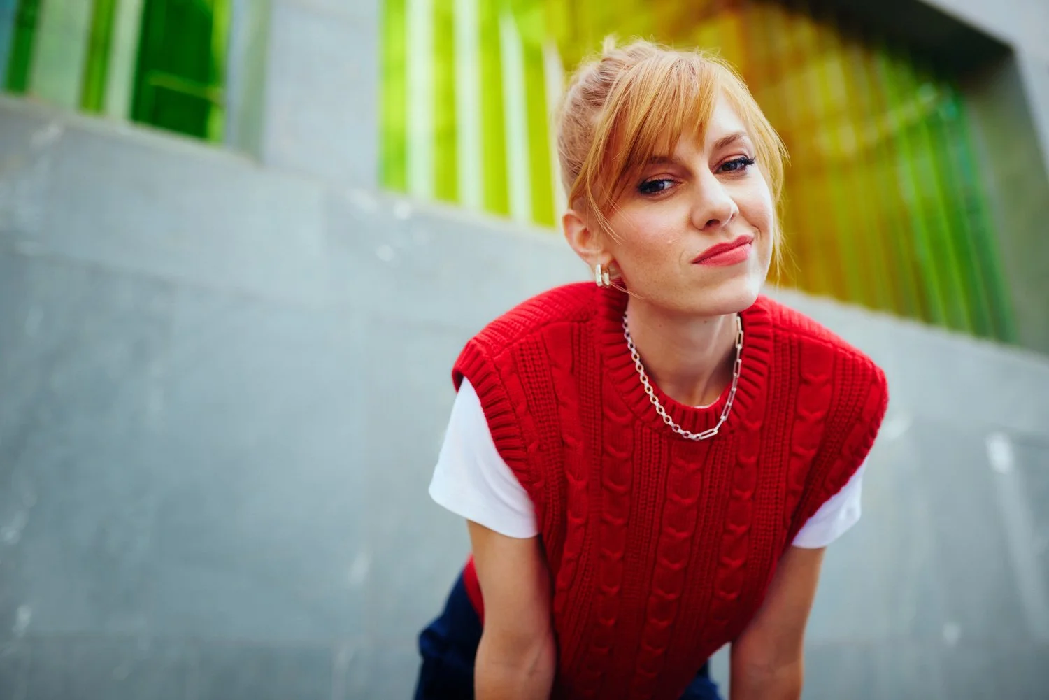 A woman with red hair, wearing a red cable-knit vest over a white shirt, standing outdoors near a concrete wall with green and yellow bars in the background.