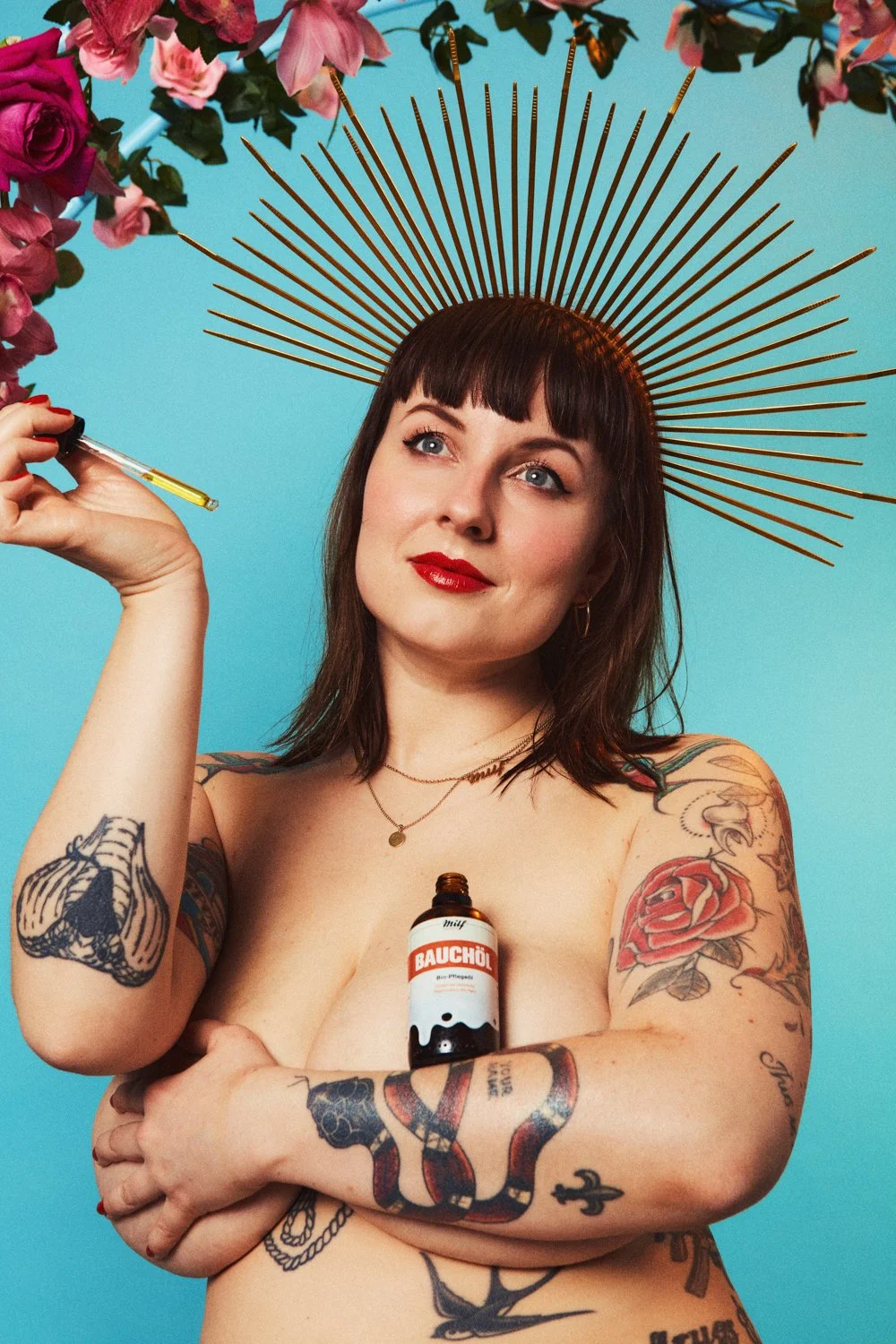 A woman with dark hair and tattoos on her arms and chest is holding a dropper and has a small bottle of Bach original flower remedy on her chest. She wears a golden halo headpiece and is standing against a bright blue background with pink flowers abo