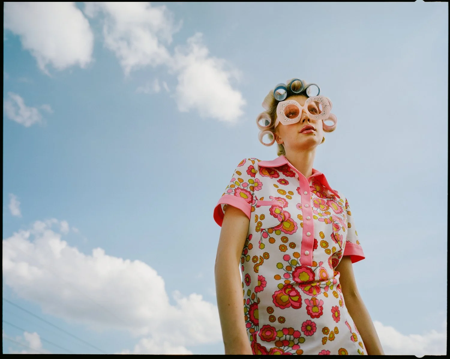 A woman wearing pink floral dress and large pink sunglasses with curlers in her hair stands outdoors against a blue sky with scattered white clouds.