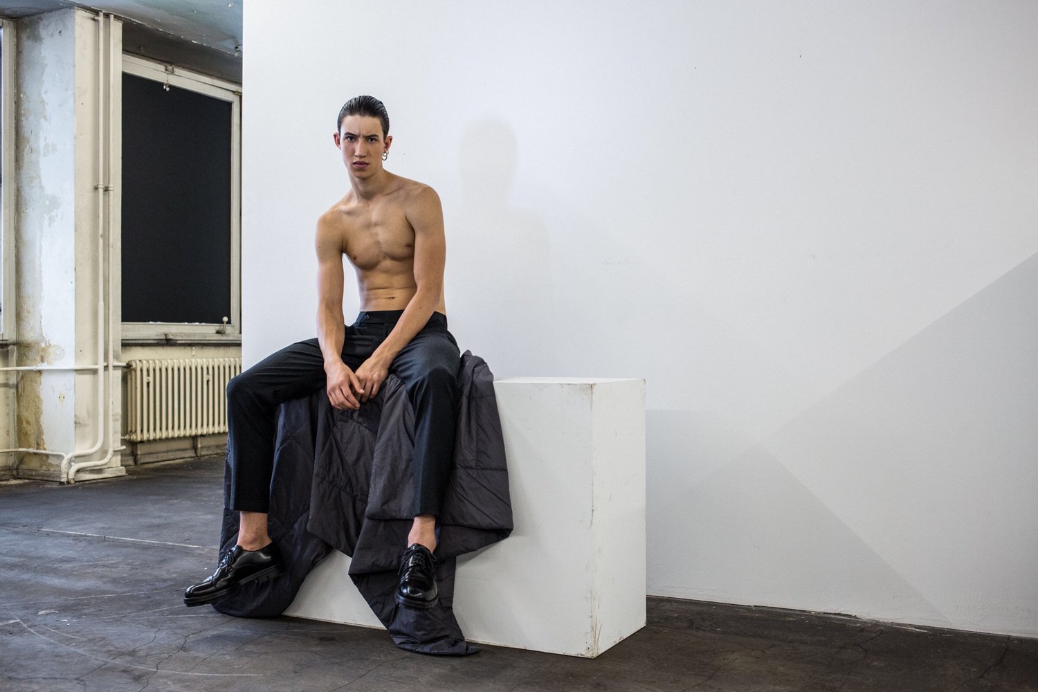 A shirtless person with dark hair sitting on a white block in a minimalist room with a black jacket and black shoes, looking at the camera.