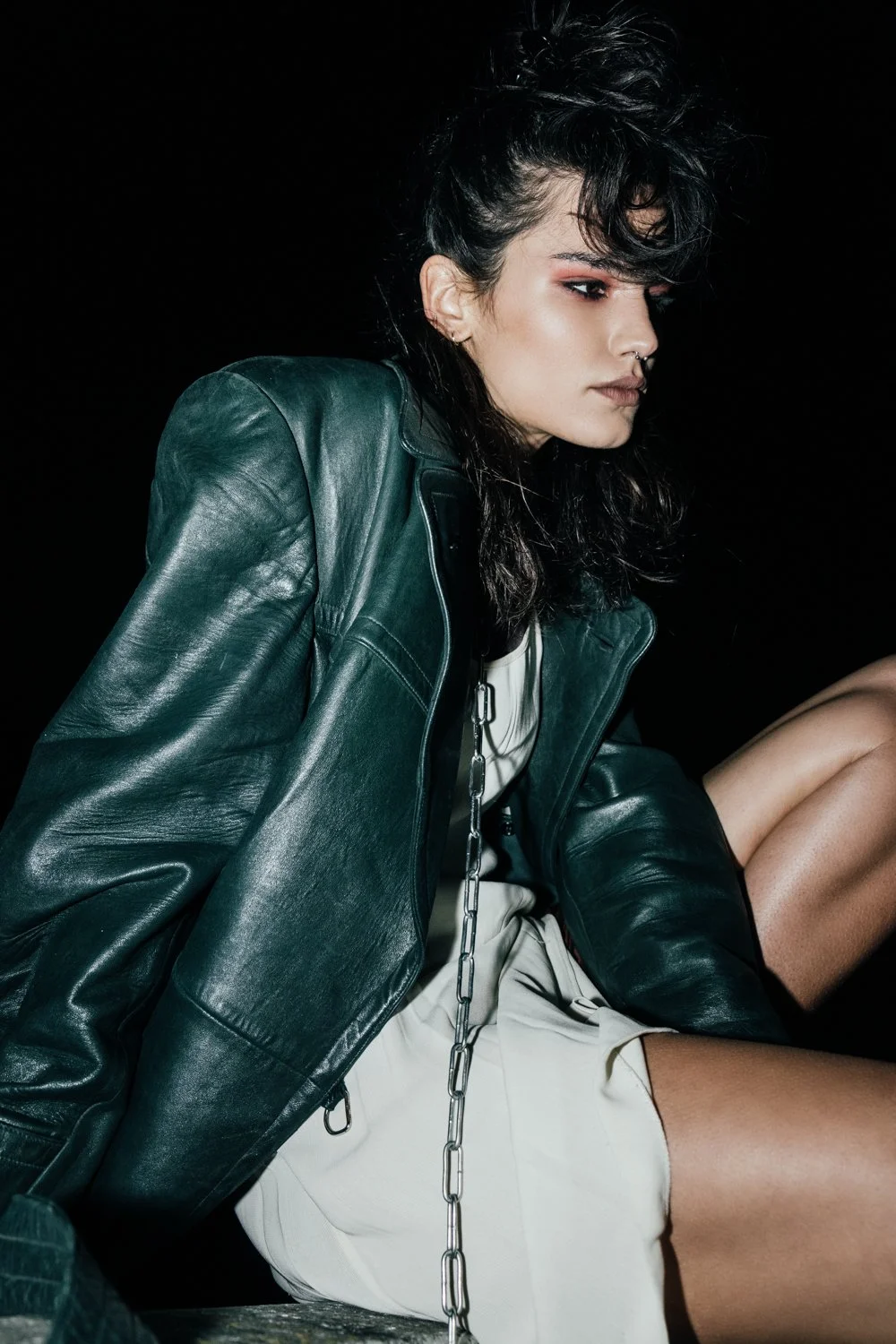 A young woman with dark, curly hair wearing a dark green leather jacket and light-colored clothing, sitting against a black background.