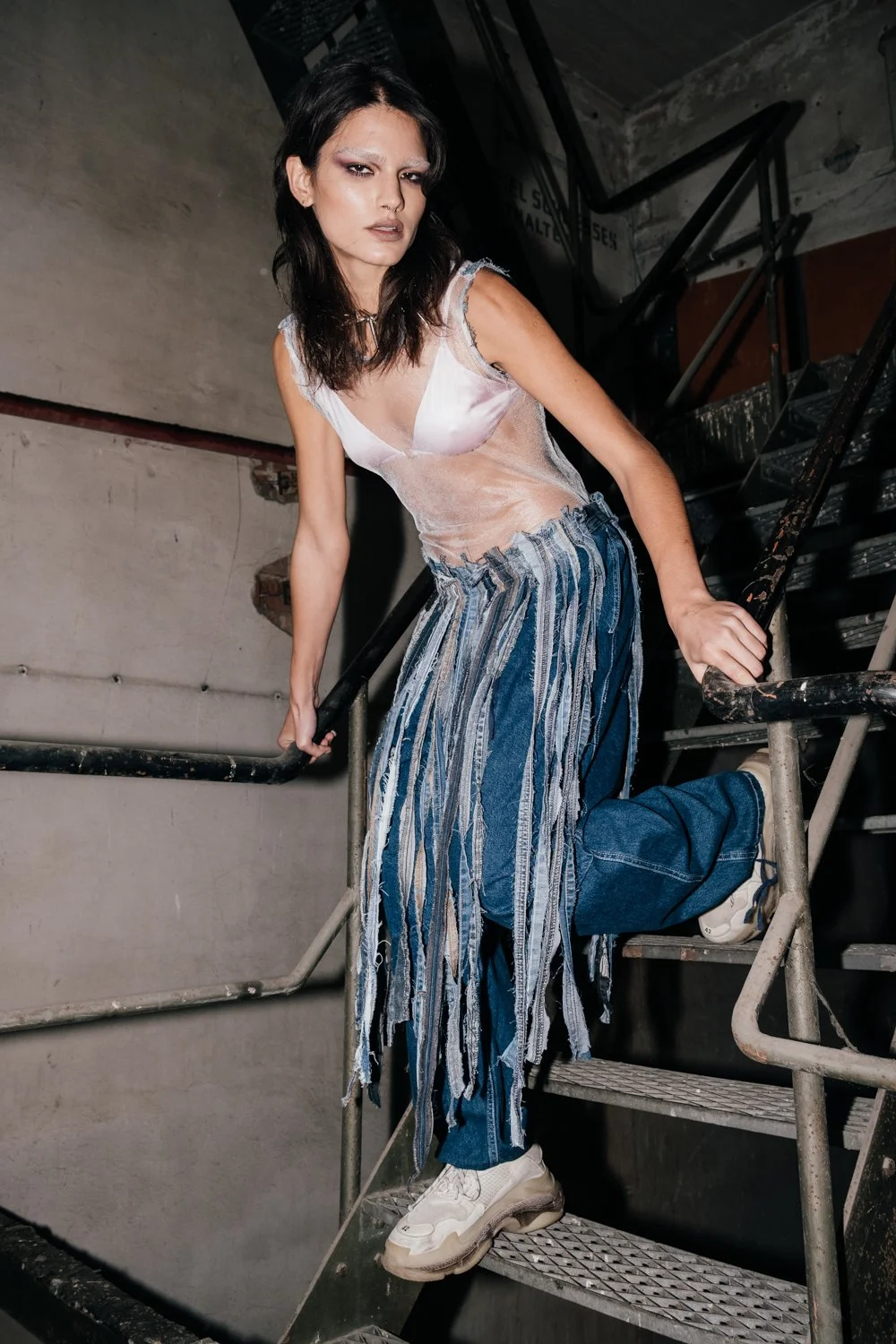 A woman with dark hair wearing a sheer or translucent top, baggy jeans with fabric strips, and sneakers, stands on a metal staircase in an industrial or urban setting.