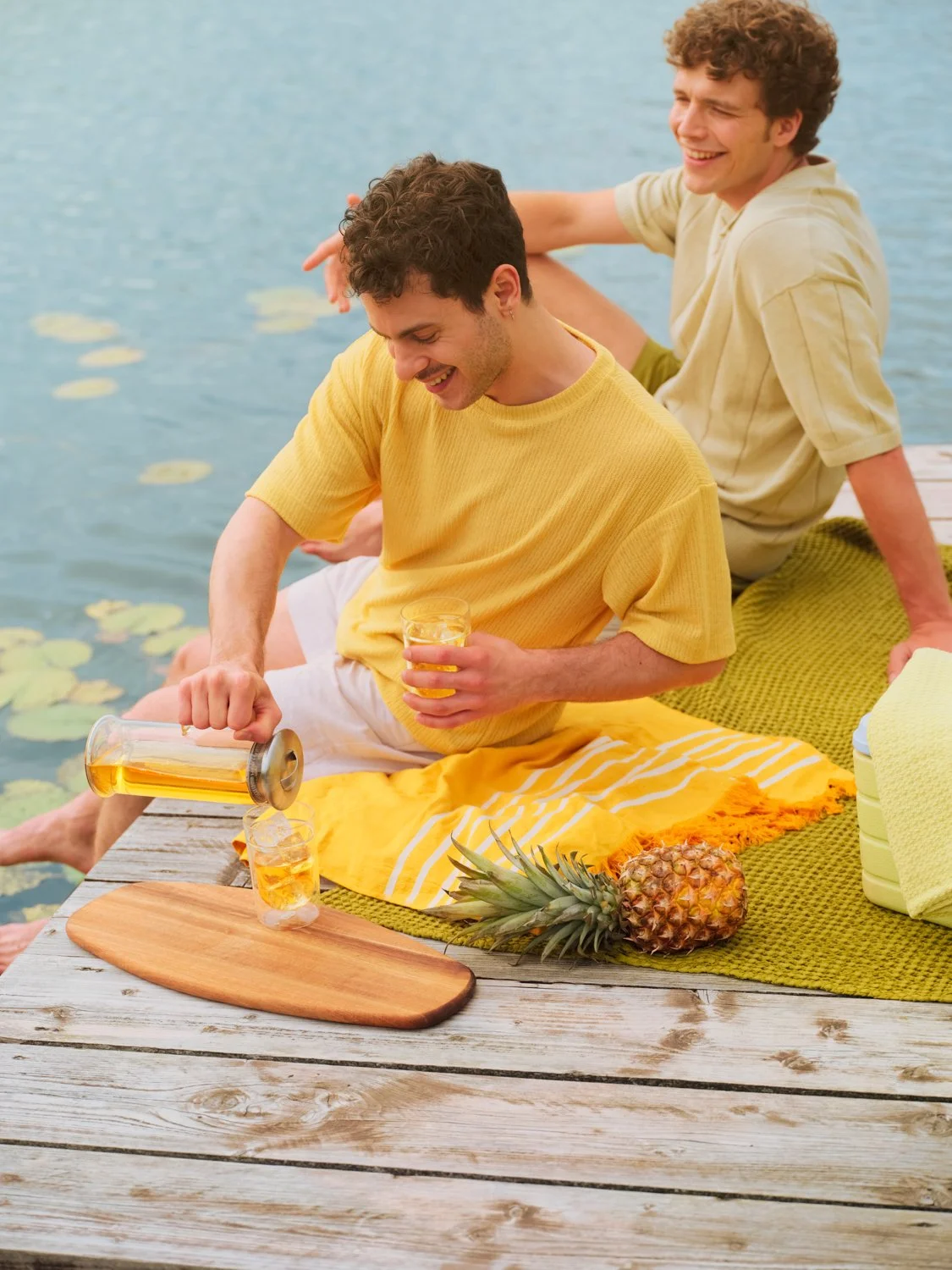 Two young men sitting on a dock by a lake, one pouring a drink into a glass, with a pineapple and other beach essentials nearby, enjoying a sunny day.