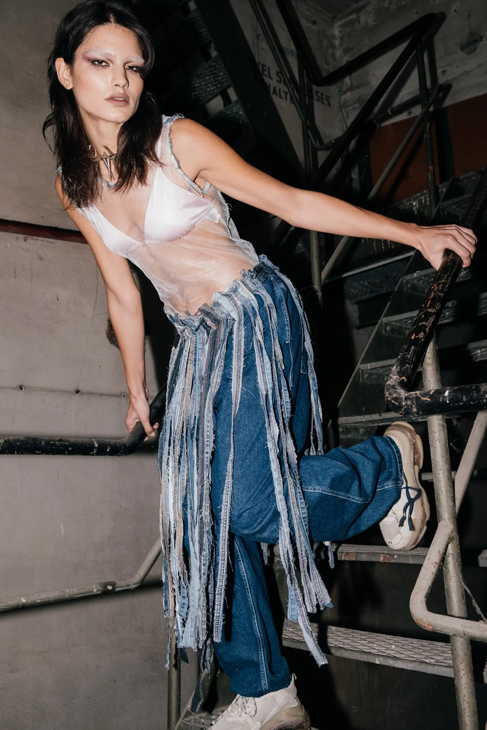A young woman with dark hair and makeup, wearing a sheer, ripped tank top and layered denim and fabric pants, stands on a metal staircase in an industrial setting.