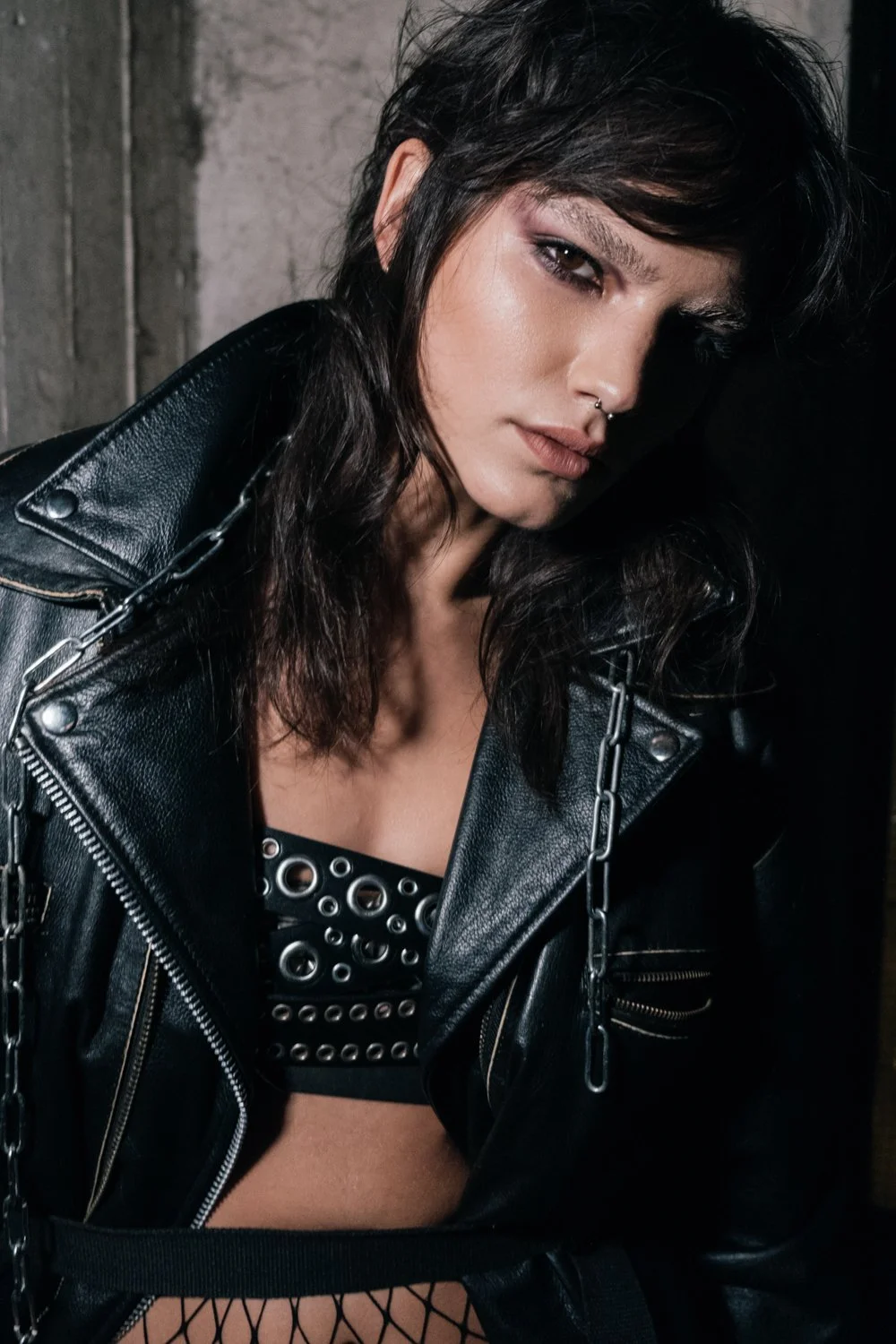 A young woman with dark hair and makeup, wearing a black leather jacket with chains, a black patterned top, and fishnet stockings, posing in a dark, industrial setting.