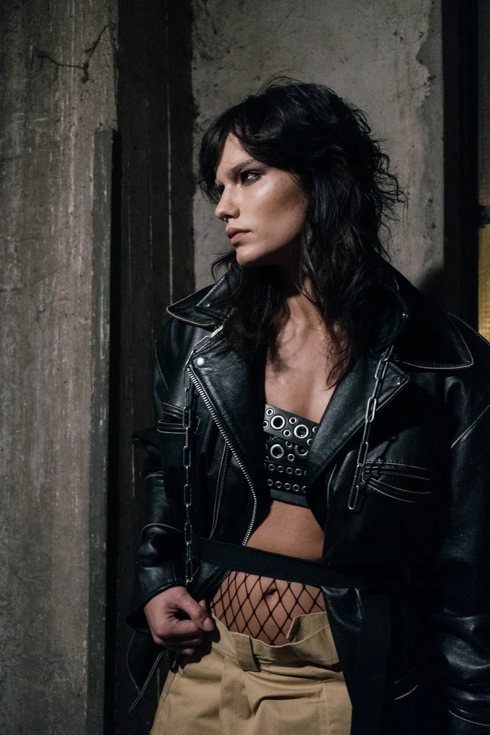 A woman with black wavy hair wearing a black leather jacket, eye makeup, fishnet stockings, and tan pants, standing against a dark, industrial wall.