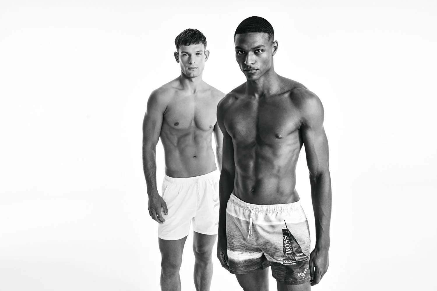 Two shirtless men wearing swim trunks against a plain white background, one in front and one behind.