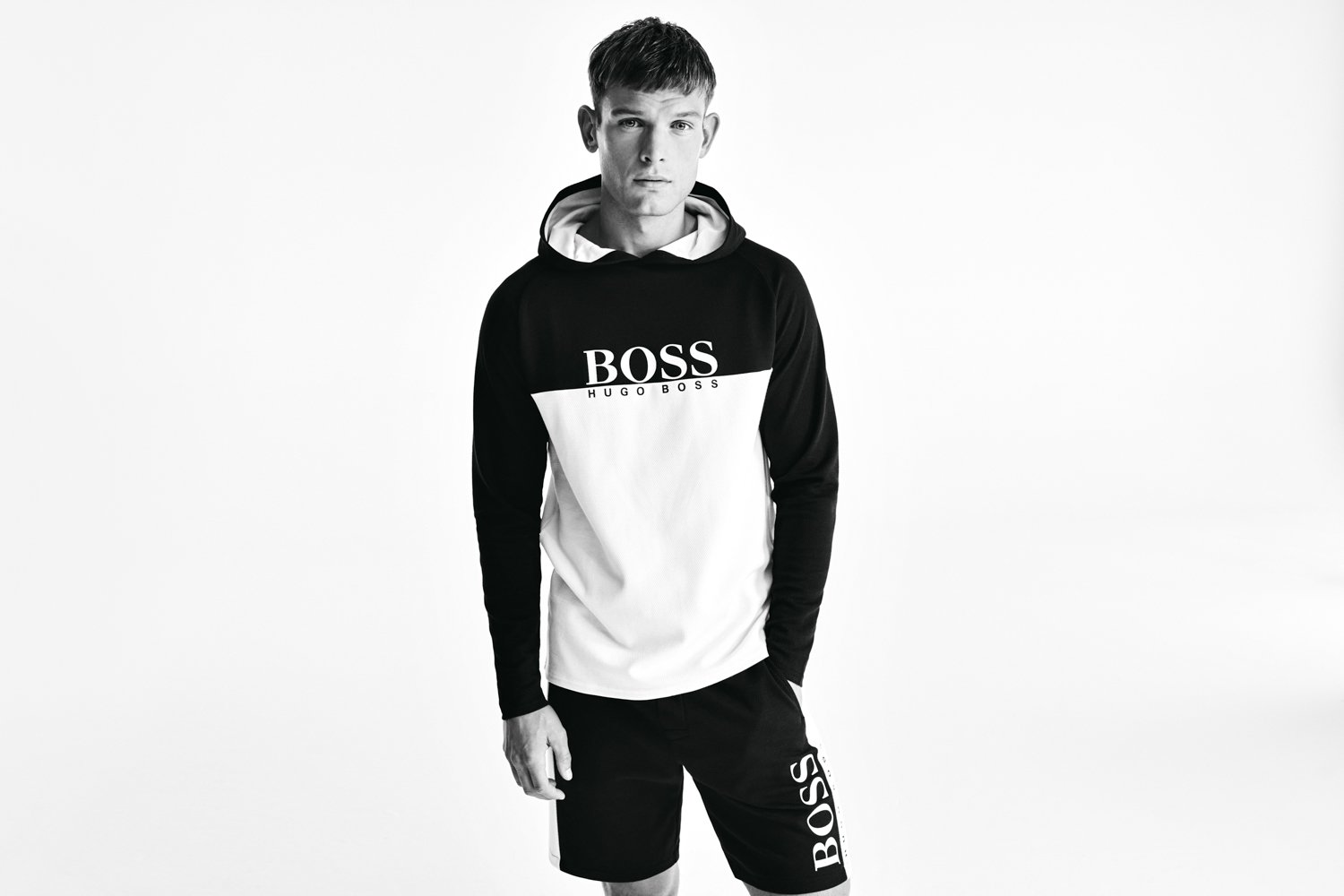 Black and white photo of a young man wearing a BOSS hoodie and matching shorts, standing with one hand in pocket against a plain white background.