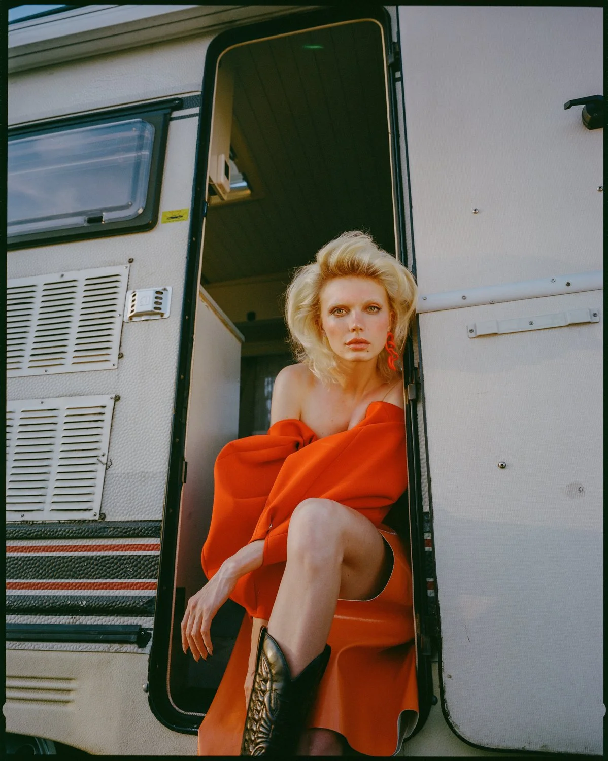 A woman with blonde hair wearing a red off-shoulder dress and black leather boots sits in the doorway of a vintage camper trailer.