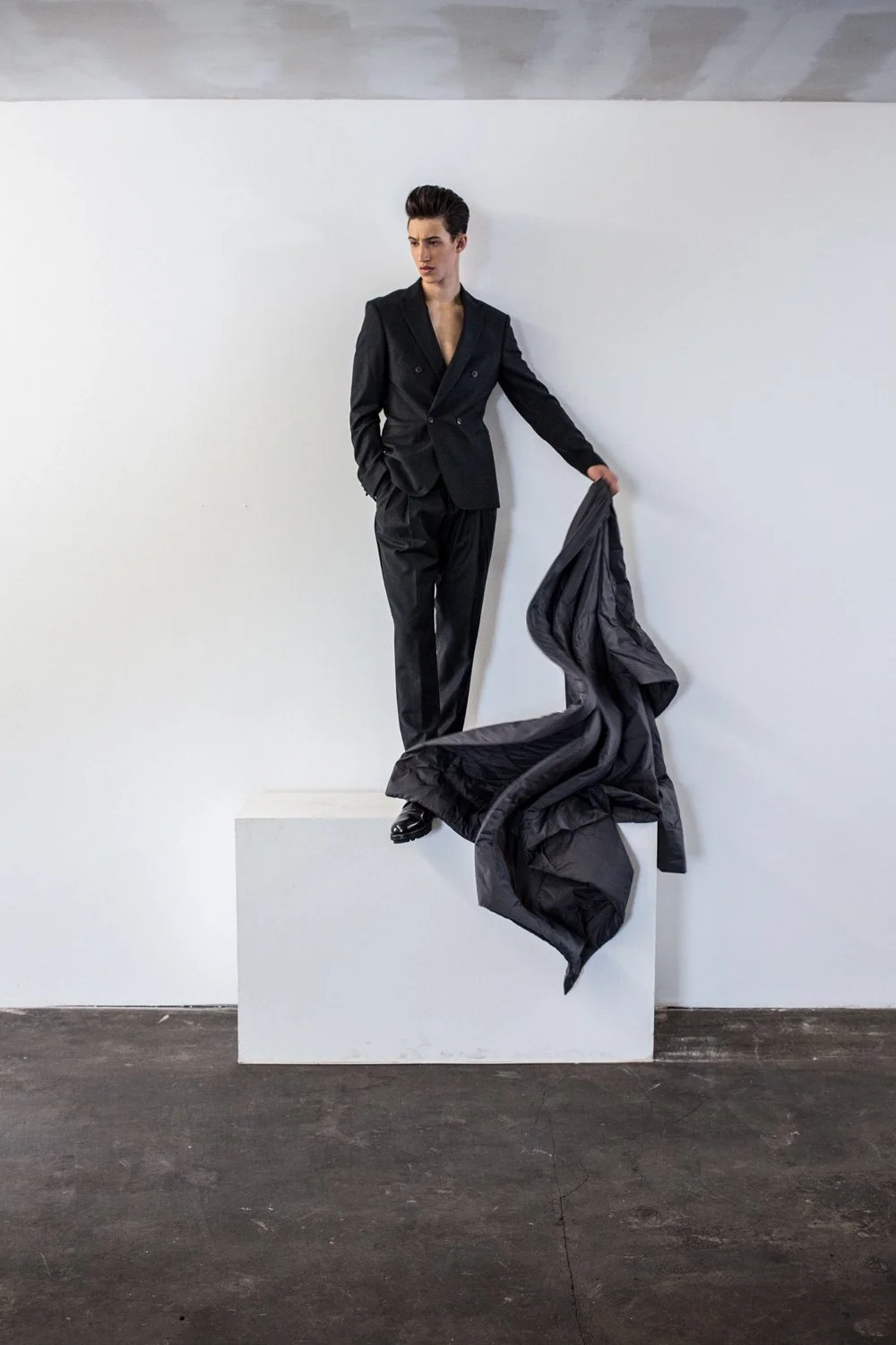 A person wearing a black suit stands on a white pedestal, holding a black jacket in one hand, with a minimalistic white wall background.