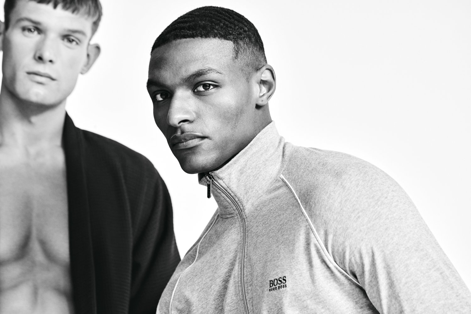 Black and white photo of three young men, one in focus with short hair and wearing a light gray BOSS sweatshirt, two others slightly blurred in the background, all with serious expressions.