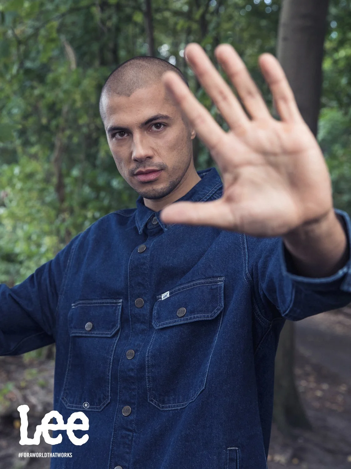 A man with a buzz cut wearing a denim shirt in a wooded outdoor setting, extending his hand toward the camera, with the Lee logo and hashtag #FORAWORLTHATWORKS in the bottom left corner.