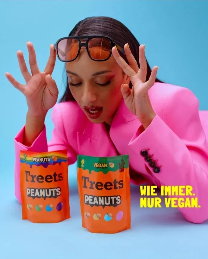 A woman wearing a pink blazer and sunglasses on her head looks at two bags of Treets Peanuts, one labeled vegan. The background is light blue, and there is yellow text in German that says, 'WIE IMMER. NUR VEGAN.'