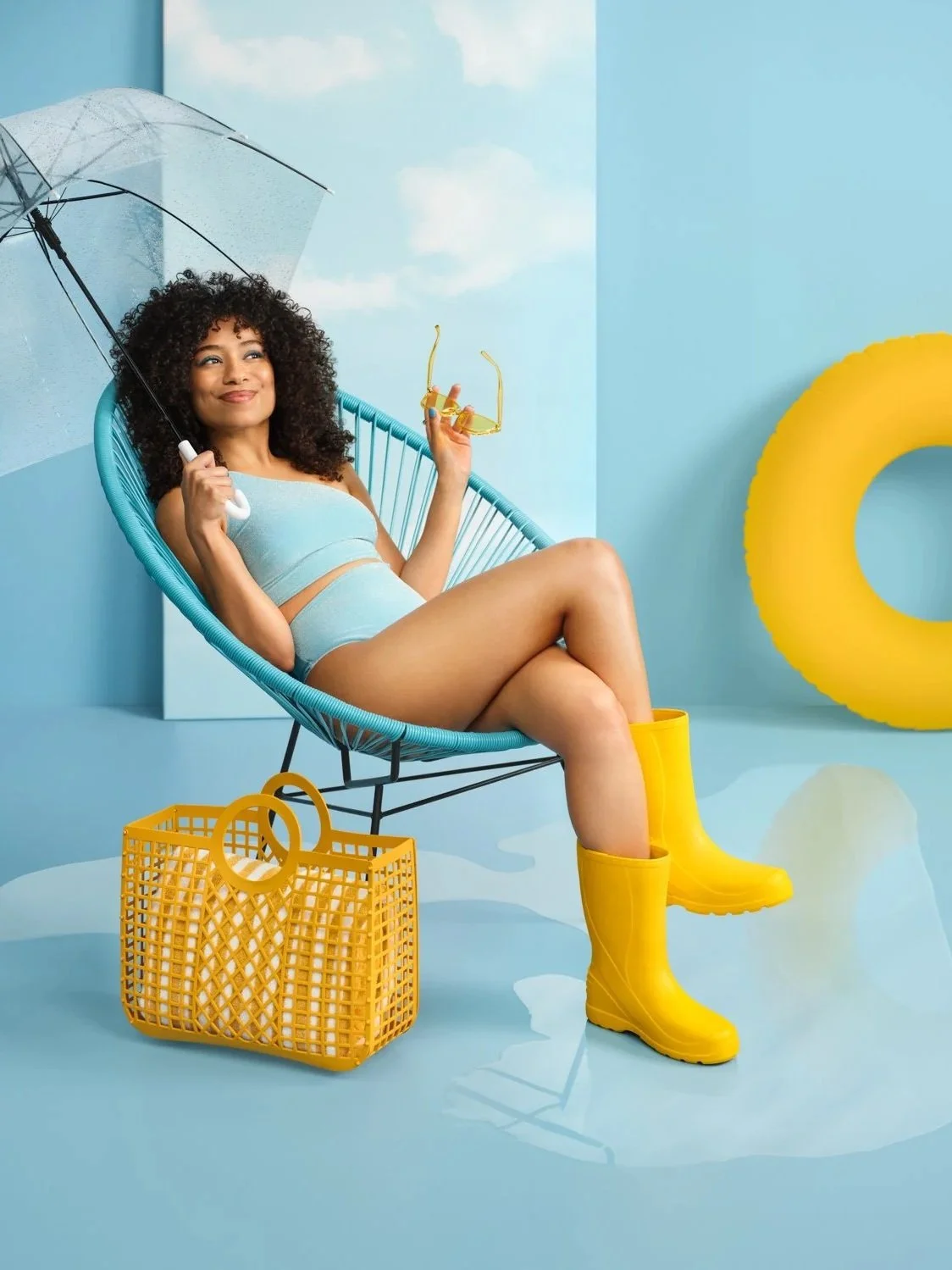 Young woman relaxing in a modern poolside setting with blue background, yellow pool float, yellow rain boots, and holding yellow sunglasses and an umbrella.