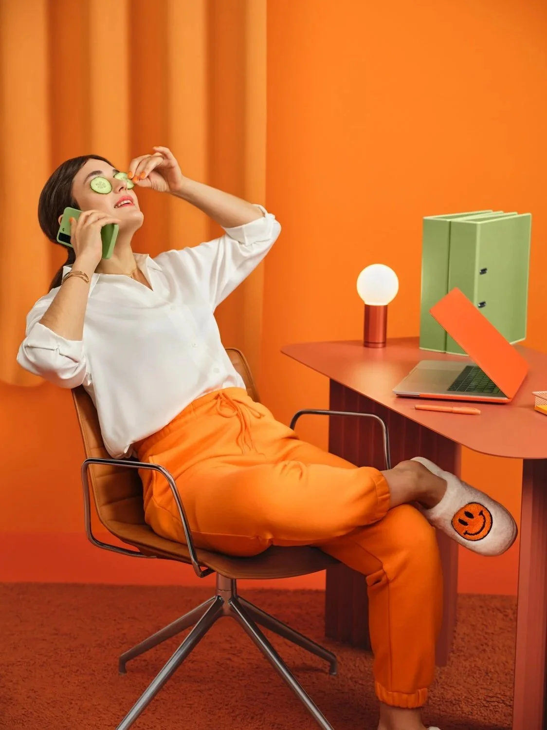 Woman sitting on an orange office chair with her feet resting on a wooden desk, wearing orange pants and gray socks with a smiley face. She has cucumber slices on her eyes and is holding a phone to her ear with one hand while nibbling on a snack with