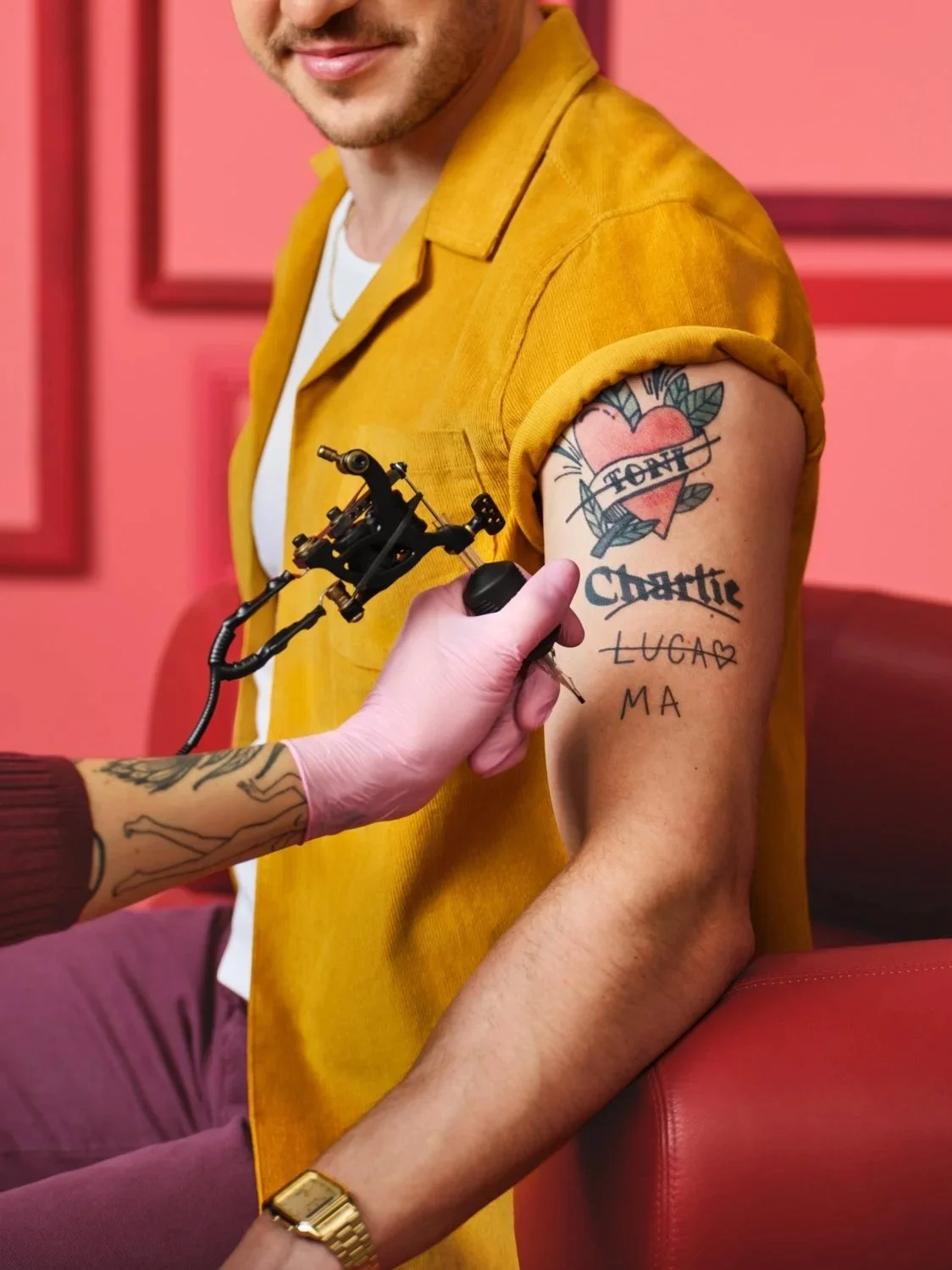 A man wearing a yellow shirt is getting a tattoo on his upper arm, which features a heart with leaves and the words 'Don't Chattie' inside the heart. The tattoo artist is using a tattoo machine, and the man's arm also has handwritten words 'LUCAD' an